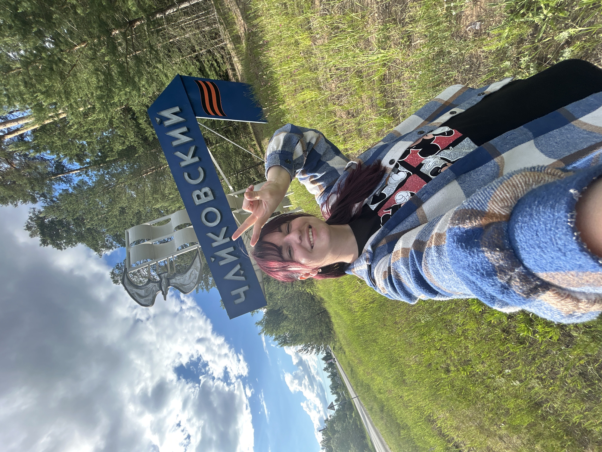A young woman with dark hair, wearing a plaid jacket over a t-shirt, smiles and makes a peace sign at the camera. She is standing in front of a large blue sign that reads "ЧАЙКОВСКИЙ" (Chaikovsky) in white Cyrillic letters, with an orange and black ribbon symbol on the right side of the sign. Behind the sign, a metal sculpture is visible against a cloudy sky. The woman and the sign are situated on a grassy slope, with pine trees visible in the background. A road can be seen in the distance, suggesting an entrance to the city. The overall scene appears to be an outdoor landmark or welcome sign.