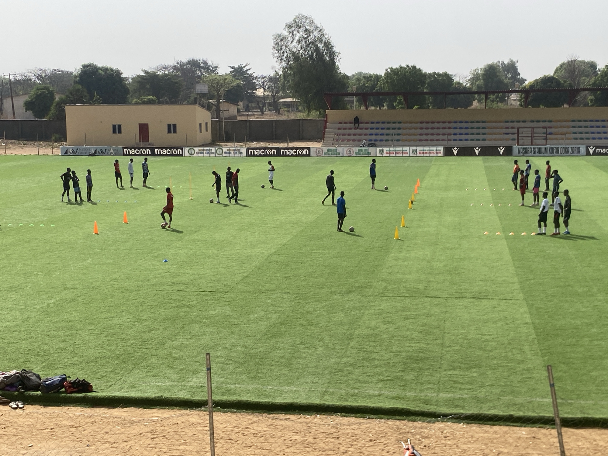 The image shows a soccer practice session at Nasara Stadium Kofar Doka Zaria, located in Tudun Wada, Nigeria. Approximately 20 players are dispersed across the green artificial turf field, some dribbling or passing soccer balls near orange and white cones. A building and stadium seating are in the background, along with a fence. Bags and shoes are visible in the foreground, near poles and the edge of the field.