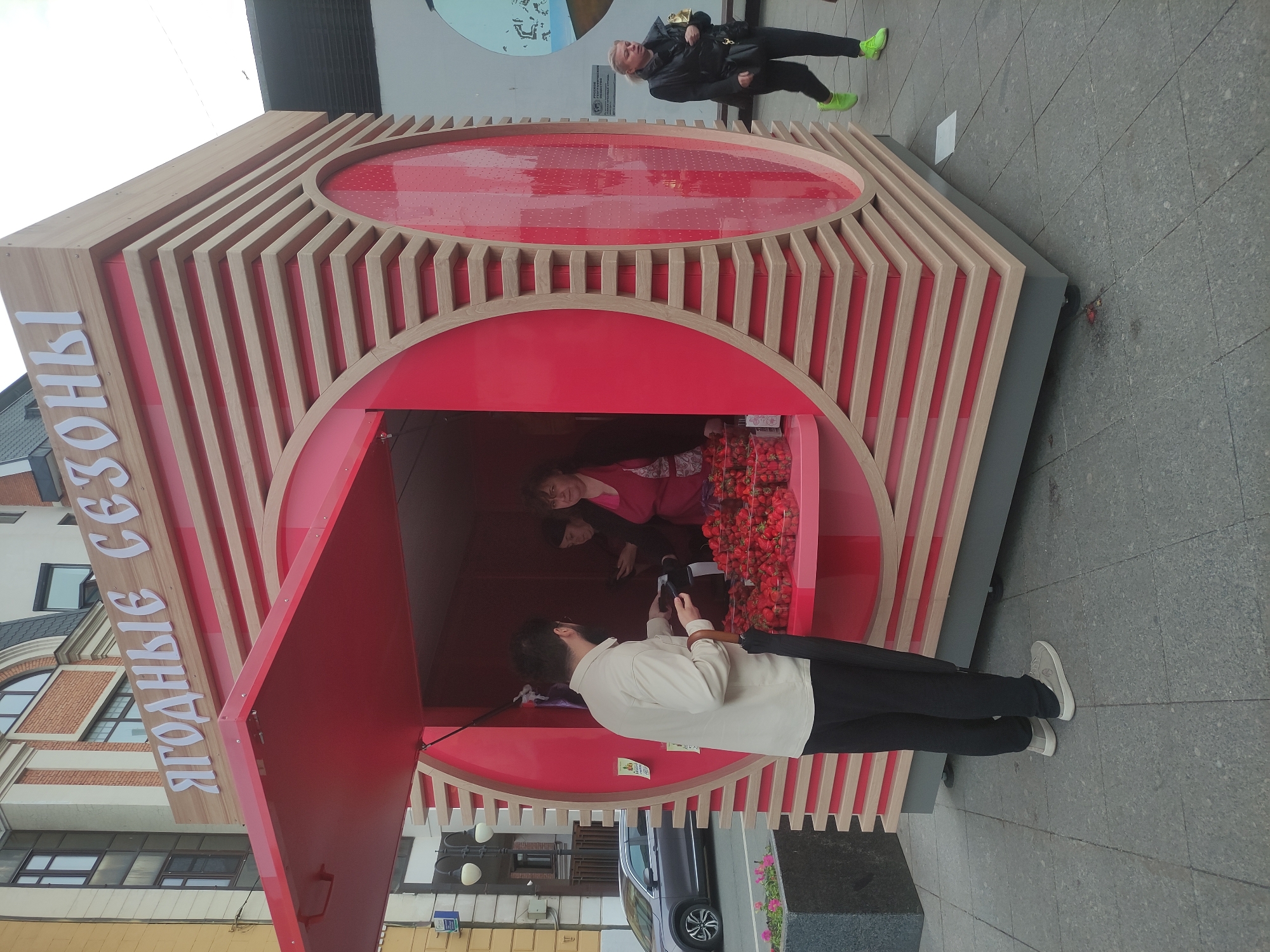 Here is a detailed description of the image:
The image depicts a small, open-air kiosk that is located in Moscow, Russia, selling strawberries. The kiosk is constructed with a frame of light wood slats interspersed with solid panels that are painted bright red, including the sales window that is open. The kiosk's name, in Russian, is written on the kiosk exterior. A person is standing outside the kiosk with an umbrella and using a smartphone pointed toward the kiosk. Inside the kiosk, two people in red outfits and strawberries are visible. A person in a black jacket is visible standing atop the kiosk. The kiosk is located on a street. Buildings can be seen in the background.