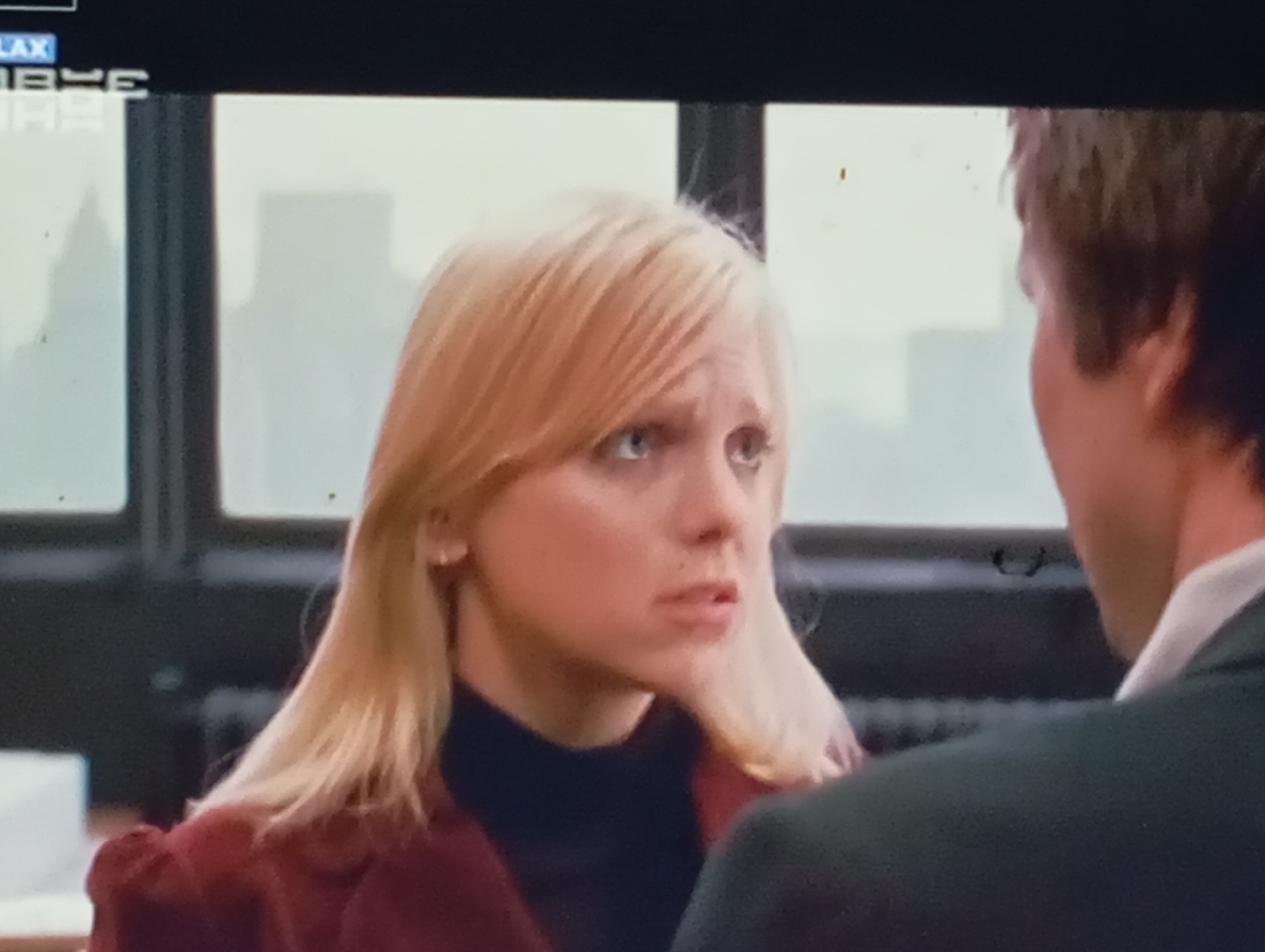 The image shows a close-up of a woman with blonde hair and a concerned expression. She is wearing a dark turtleneck and a burgundy jacket. Her head is turned to her right, looking at someone or something outside the frame. The background appears to be an indoor setting with a window showing a blurred cityscape. The woman's makeup is visible, and she has light-colored eyes. The image is slightly grainy, suggesting it might be a still from a video or film. There is no indication of the city of Sofiivska Borshchahivka, Ukraine, or any specific geographical location. The scene appears to be a candid moment captured in an urban environment.