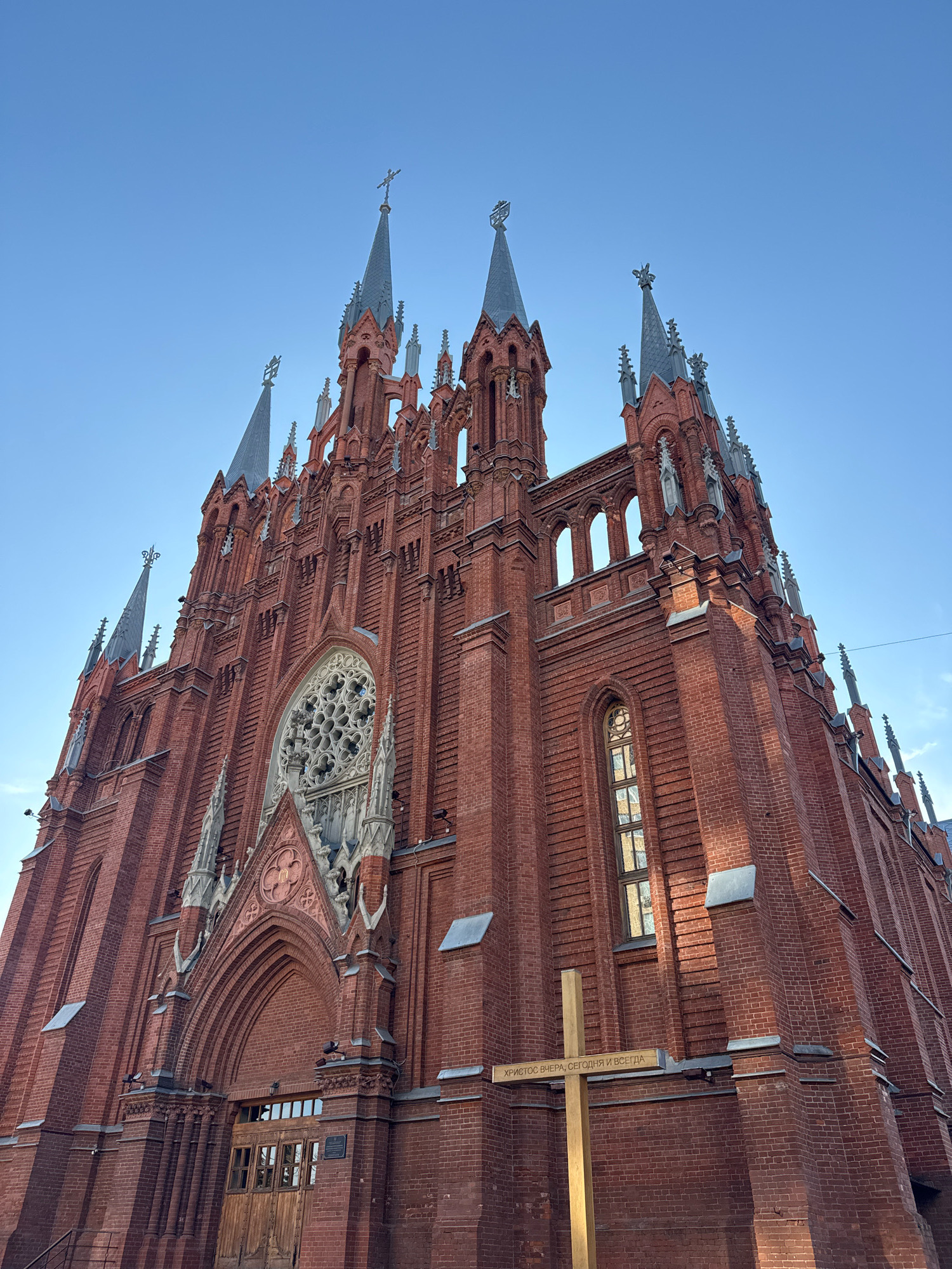 The image shows a low-angle view of the Cathedral of the Immaculate Conception in Moscow, Russia. The Gothic Revival cathedral, constructed from red brick, features multiple spires, a rose window above the entrance, and arched windows. A large wooden cross stands in front of the cathedral, bearing an inscription in Russian that translates to "Christ yesterday, today, and forever." The sky is clear and blue.