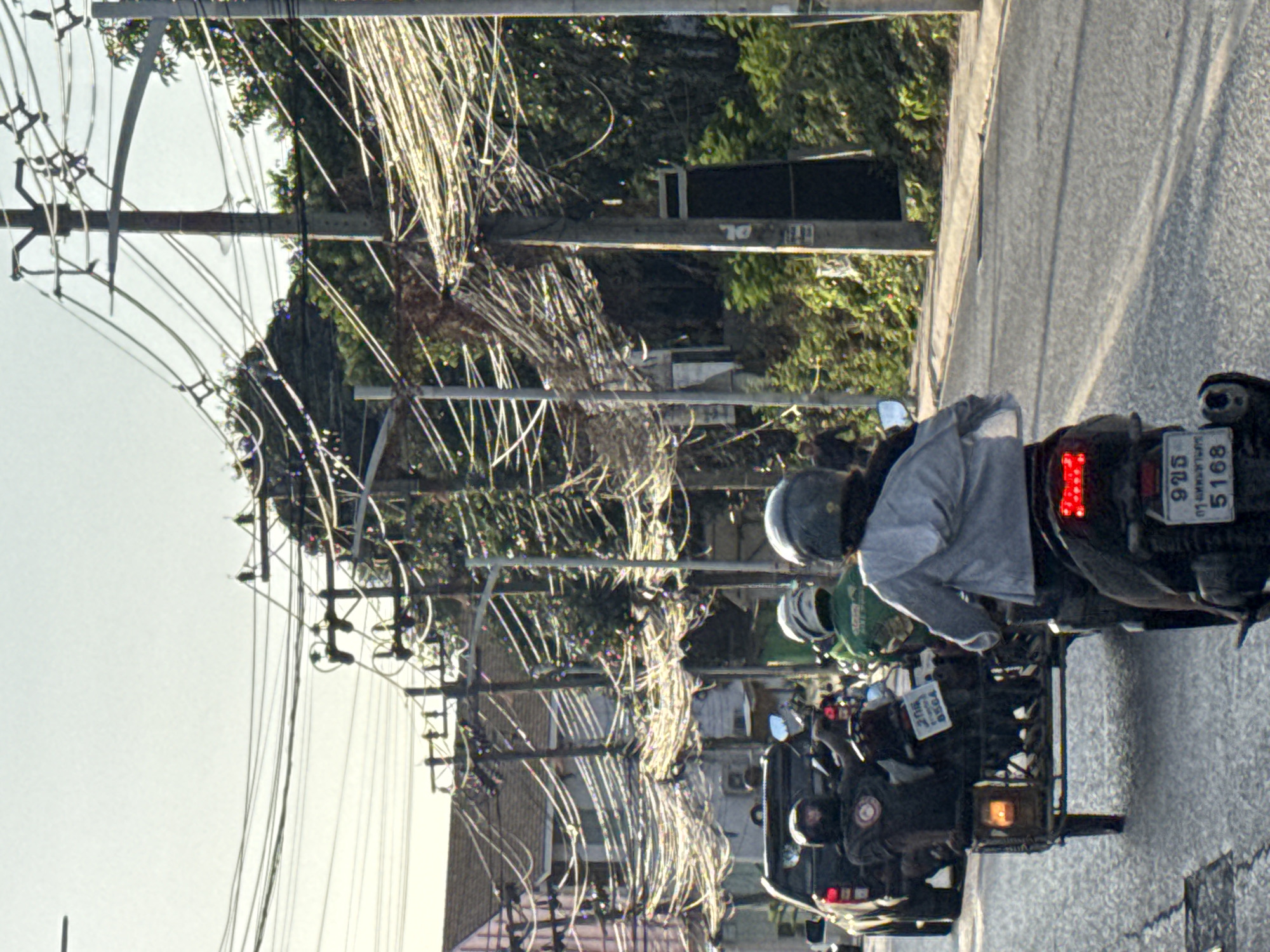 The image depicts a street scene in Ko Samui District, Thailand, characterized by a dense network of overhead electrical wires. In the foreground, a person is riding a motorcycle, their back to the camera, wearing a gray hooded jacket and a helmet. The motorcycle has a red taillight and a license plate with Thai characters and the number "5168". To the left, a motorcycle with a sidecar is moving, carrying two individuals also wearing helmets. The driver in the sidecar is wearing a dark jacket with a circular emblem, and the passenger is wearing a green helmet. The sidecar motorcycle has a license plate with Thai characters and the number "6564". Further down the street, a dark-colored pickup truck is visible. The environment features buildings with tiled roofs on the left side of the road and lush green vegetation on the right. Utility poles with numerous tangled wires dominate the upper portion of the frame, suggesting a cluttered urban or semi-urban setting. The sky is bright and appears to be daytime.
