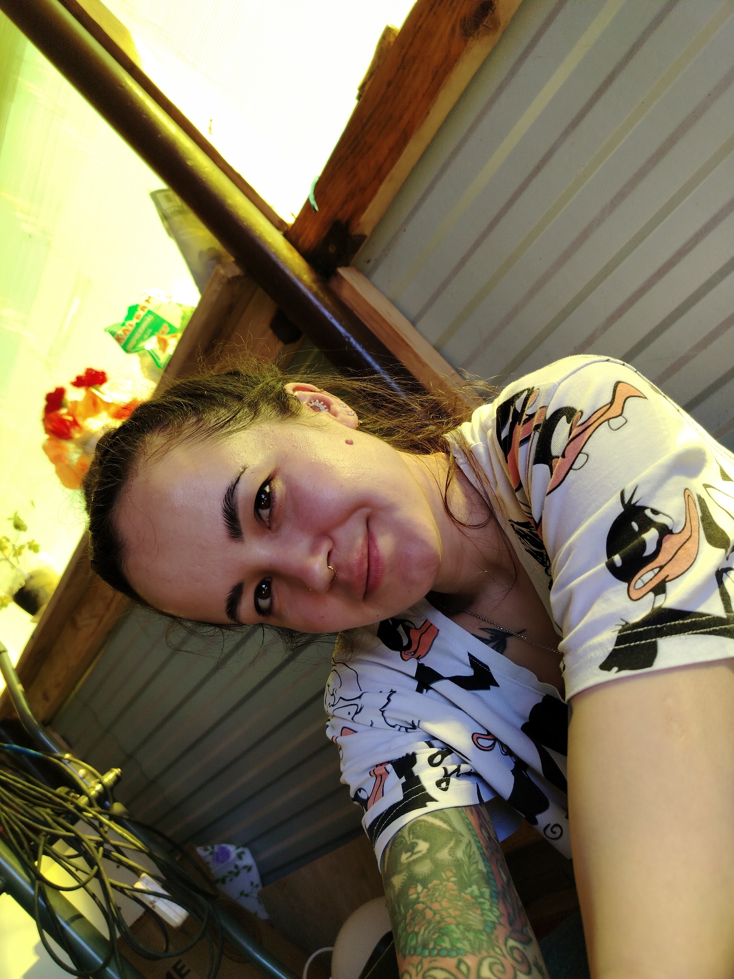 A woman with dark hair pulled back, a nose ring, a mole on her right cheek, and multiple ear piercings poses for a selfie. She is wearing a white t-shirt with a cartoon duck pattern and has a colorful tattoo on her left arm. The location appears to be inside a structure with a corrugated metal roof and wooden beams. Electrical cables are visible in the lower left corner. The location may be in Гожан, Russia.