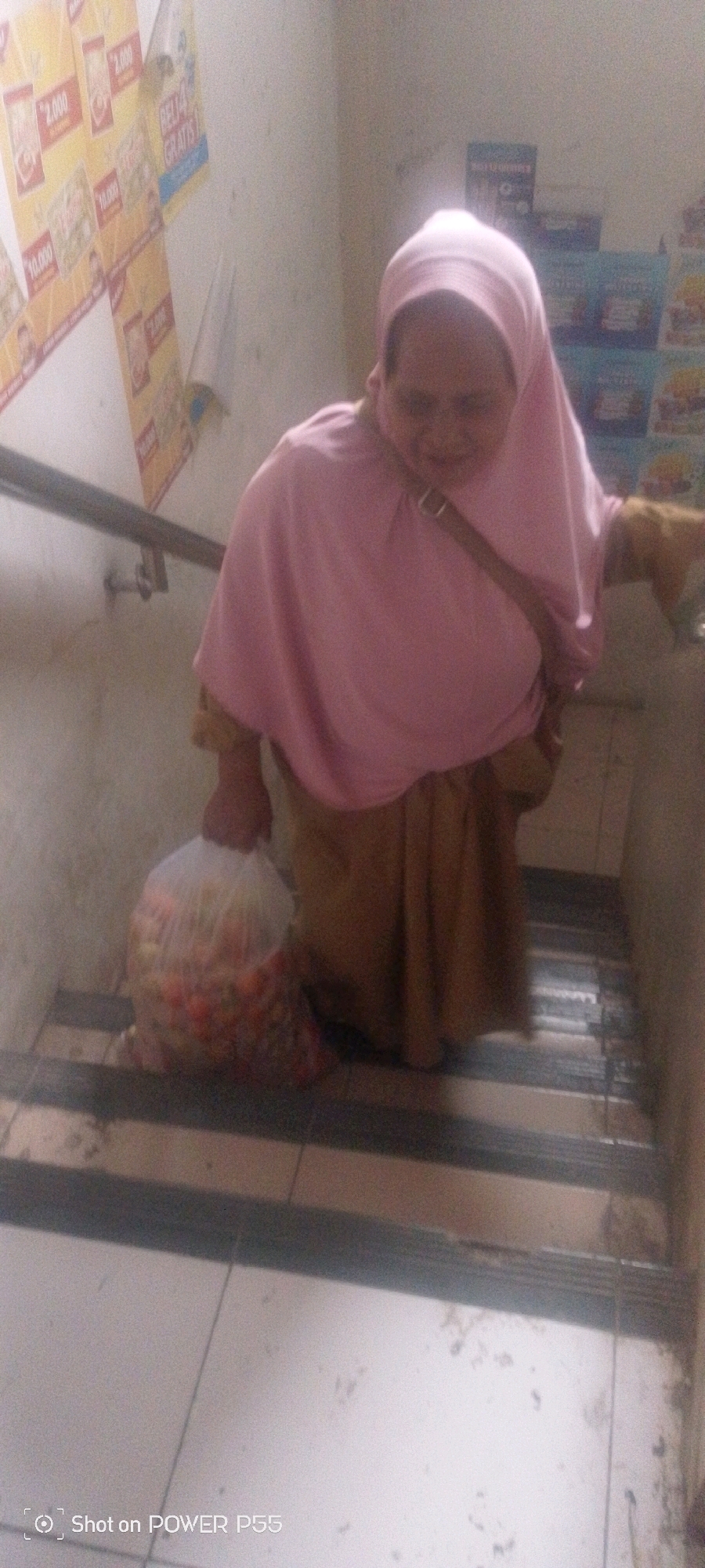 An individual wearing a pink hijab and a brown skirt is walking up a flight of stairs while carrying a large plastic bag filled with produce. The person is looking down and appears to be focused on their descent. The stairs are made of concrete and have a metal handrail on the left side. The walls are painted a light color and are adorned with posters that are difficult to read. The lighting is dim, and the overall atmosphere of the scene is unremarkable.