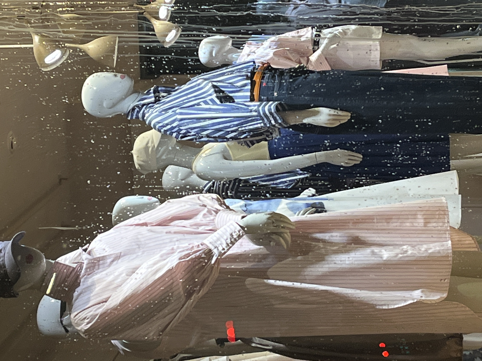 The image depicts a window display with several mannequins. The mannequins are arranged horizontally, one above the other, and dressed in casual clothing. The clothing includes striped shirts, jeans, and other garments. The mannequins have plain white heads and are positioned to appear as though they are lying down. The display is behind glass, which has water droplets or other imperfections that distort the view slightly. There are also ceiling lights visible above the mannequins. Without further information, the specific location in Krasnoyarsk, Russia cannot be determined.