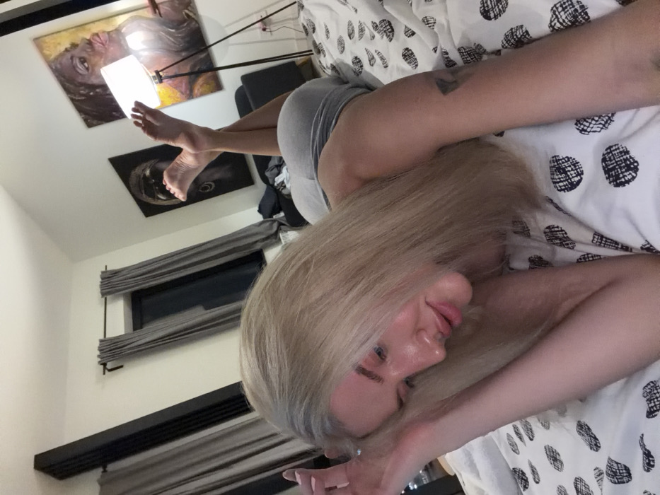 A woman with blonde hair rests on a bed with a patterned duvet cover. She wears grey shorts and has a tattoo on her arm. Above her, a floor lamp illuminates her feet, which are raised towards the ceiling. Two pieces of art hang on the wall, depicting faces. A window with grey curtains is also visible. The image is taken at a tilted angle.