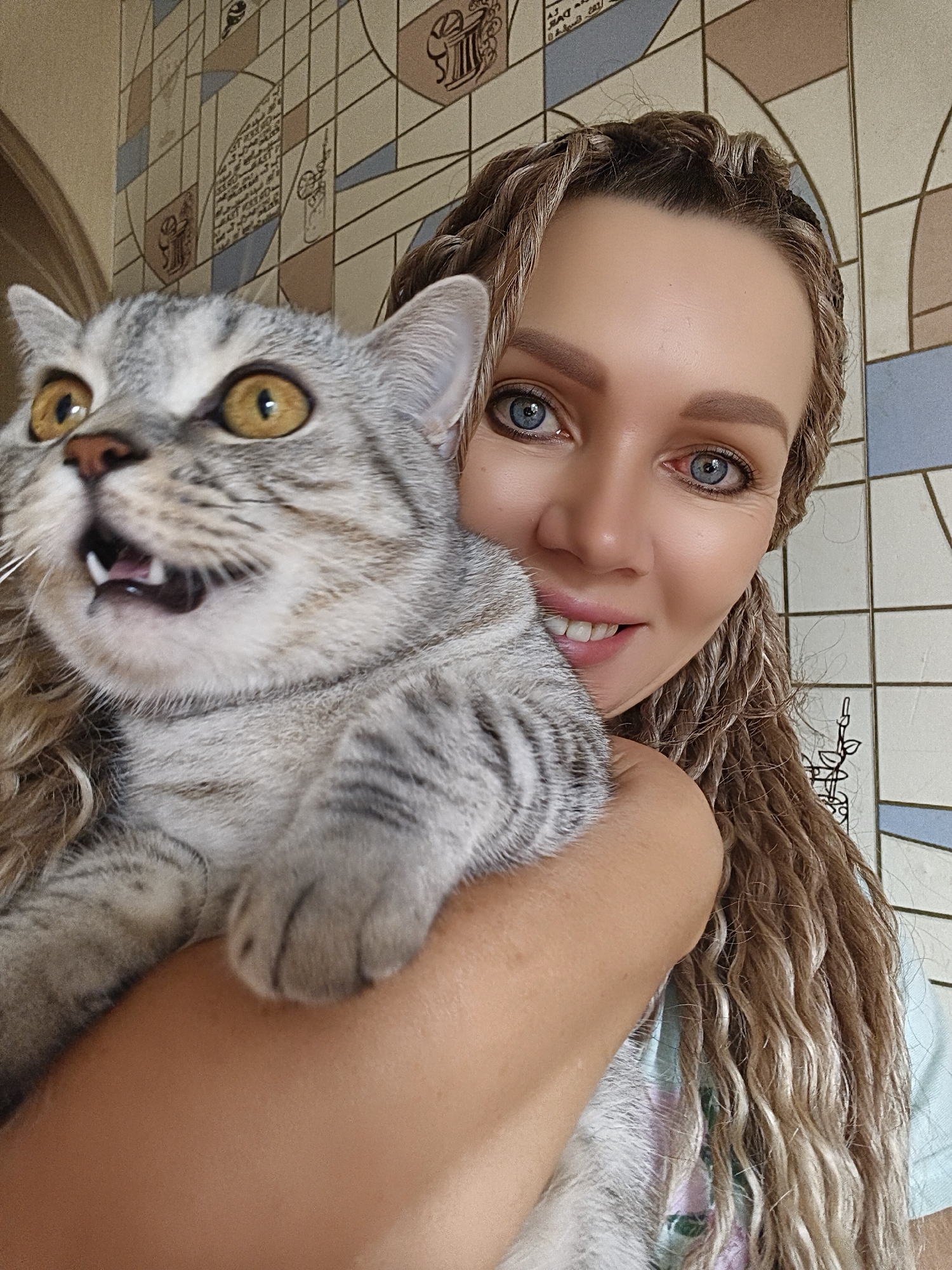 A young woman with blonde braided hair and blue eyes smiles at the camera, holding a grey tabby cat. The cat is looking off to the side with its mouth open, revealing its teeth. The woman is wearing a light blue shirt. They are in front of a wall with a tiled pattern. The scene appears to be indoors.