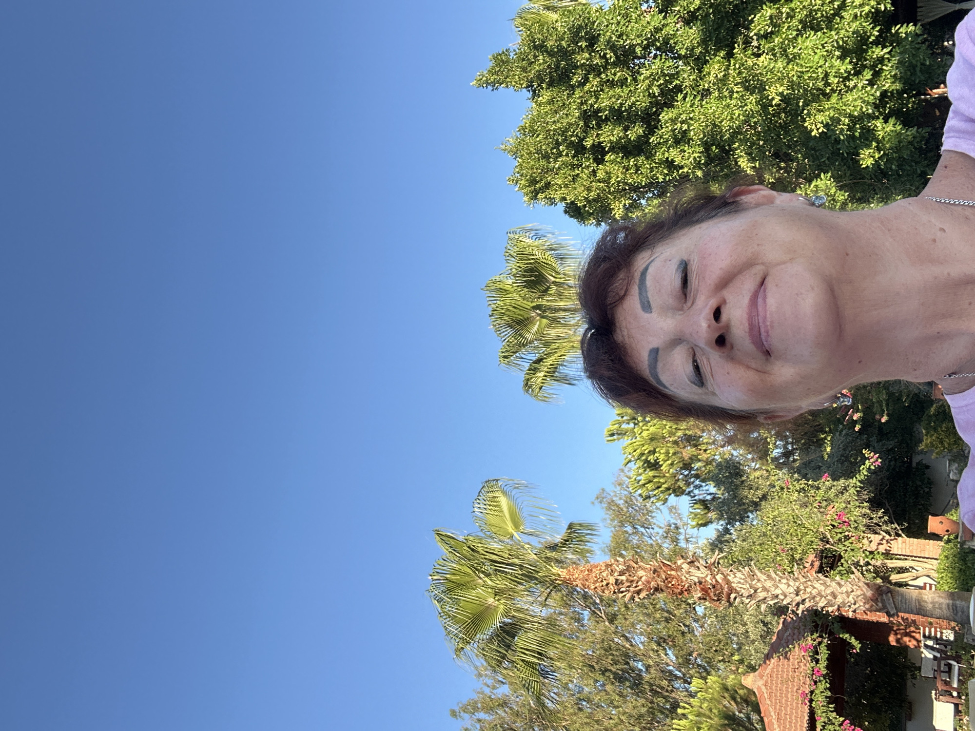 An outdoor selfie is taken by a woman with auburn hair and defined eyebrows. She is smiling with her eyes closed and is positioned in the foreground, occupying the lower portion of the frame. Behind her, a vibrant blue sky dominates the upper two-thirds of the image. The mid-ground reveals lush green foliage, including several palm trees and other dense trees. To the left, a small, reddish-brown gazebo with flowering pink vines is visible, along with outdoor furniture. The scene appears to be set in a warm, sunny environment, likely a garden or resort area. The setting is consistent with the region of Ortaca, Turkey, known for its Mediterranean climate and abundant greenery.