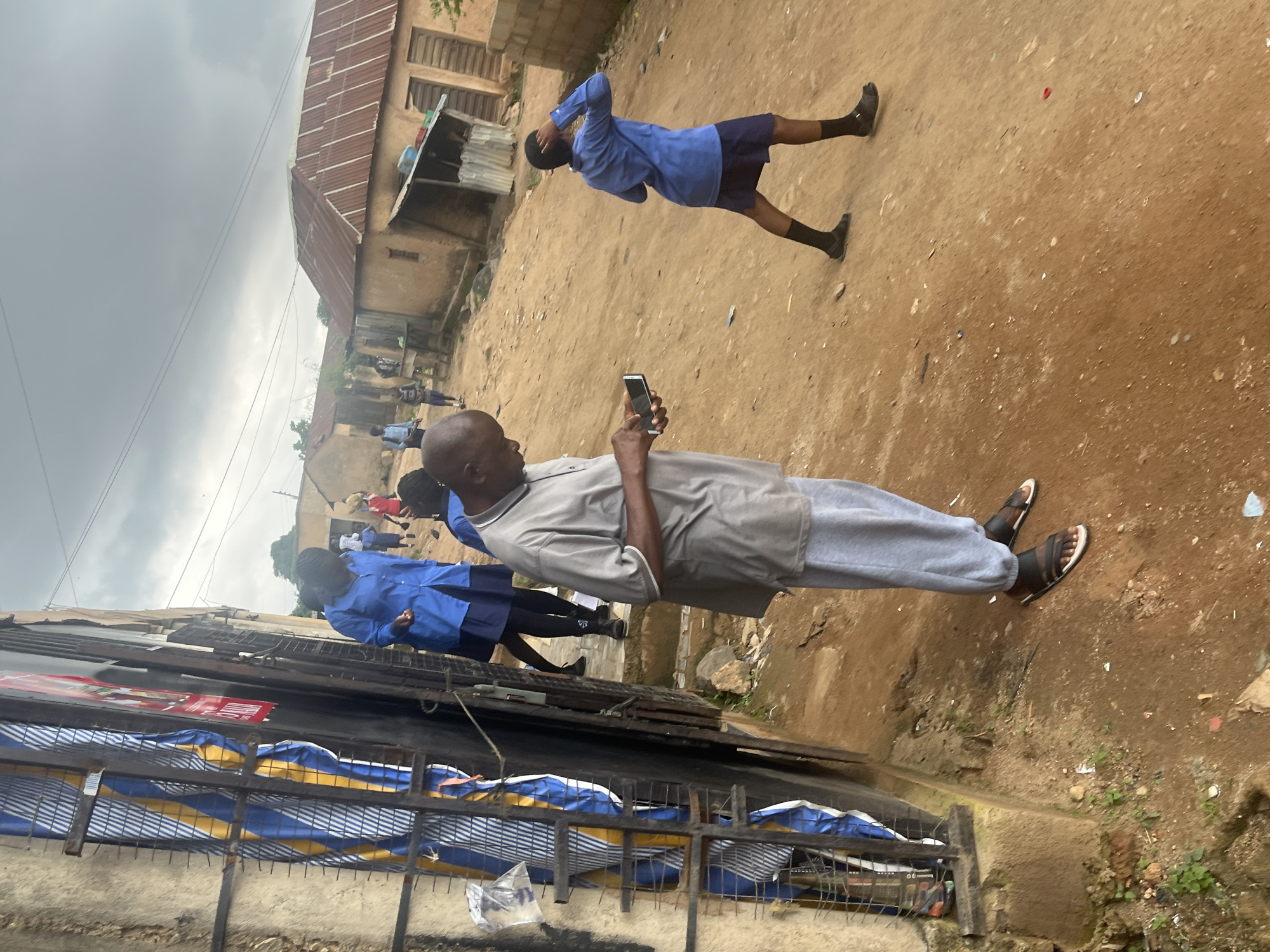 The image depicts a street scene in Gura Top, Nigeria. In the foreground, a man in a grey polo shirt and light-colored pants is looking at his smartphone. He is wearing sandals. Behind him, several young people in blue uniforms, possibly students, are also present. One student is captured in motion, appearing to be performing an athletic move. The ground is a dusty, dirt surface, with some debris scattered around. In the background, there are simple buildings with corrugated metal roofs, characteristic of many informal settlements. The sky is overcast, suggesting a cloudy day, possibly before or after rain. The overall impression is one of everyday life in a community where infrastructure appears basic. There are no discernible signs with legible text. The image seems to be taken during daylight hours.