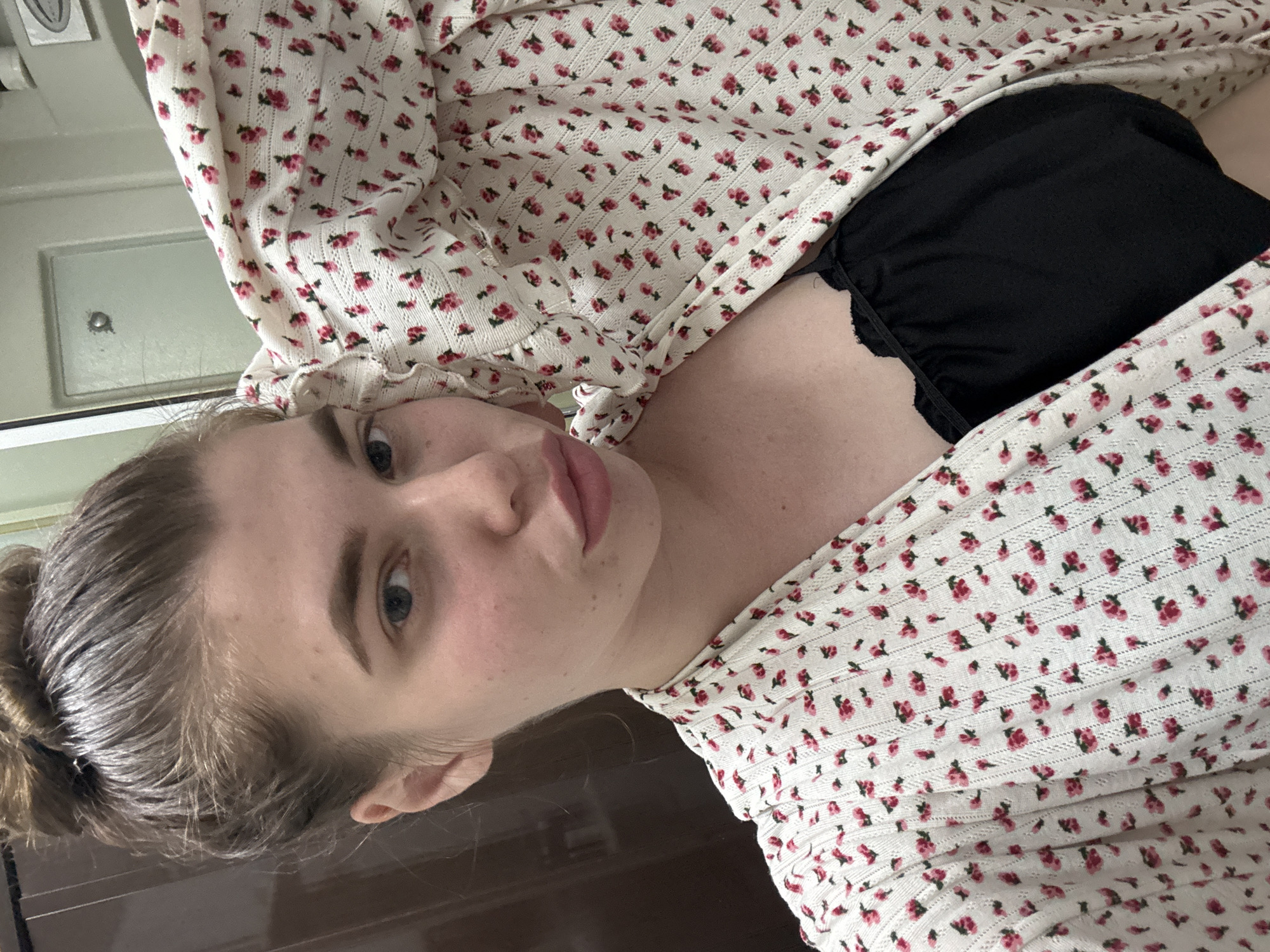 A woman with fair skin and blue eyes is lying down. She has her hair pulled back in a bun. She is wearing a white floral top over a black garment, possibly a bra or camisole. The background is blurry, but there is a door visible in the upper left corner and a brown surface beneath her. The image does not provide any information regarding the city of Мещёры, Russia.