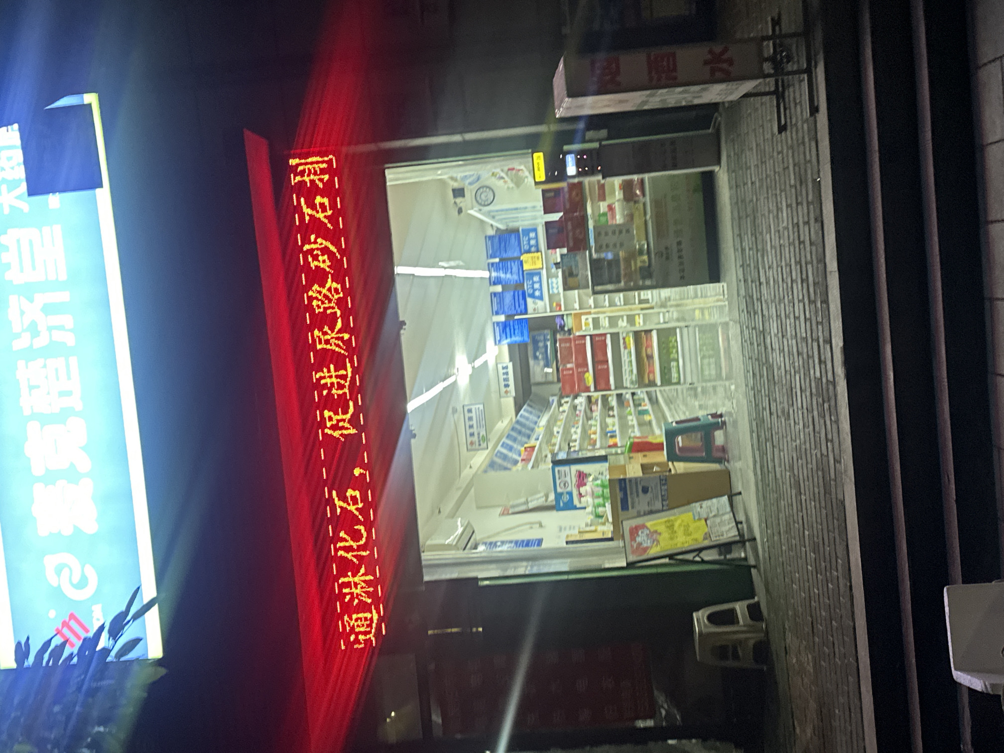 The image shows the exterior of a pharmacy at night. A bright blue and yellow neon sign is visible at the top, with Chinese characters. Below this, a red neon sign displays more Chinese characters. The pharmacy's entrance is wide and well-lit, revealing shelves stocked with various products. Several white and green plastic stools are placed outside the entrance. A red and white sign with Chinese characters is visible on the right side of the image. The ground in front of the pharmacy is paved with bricks. The location is identified as Chongqing High-tech Zone, China.