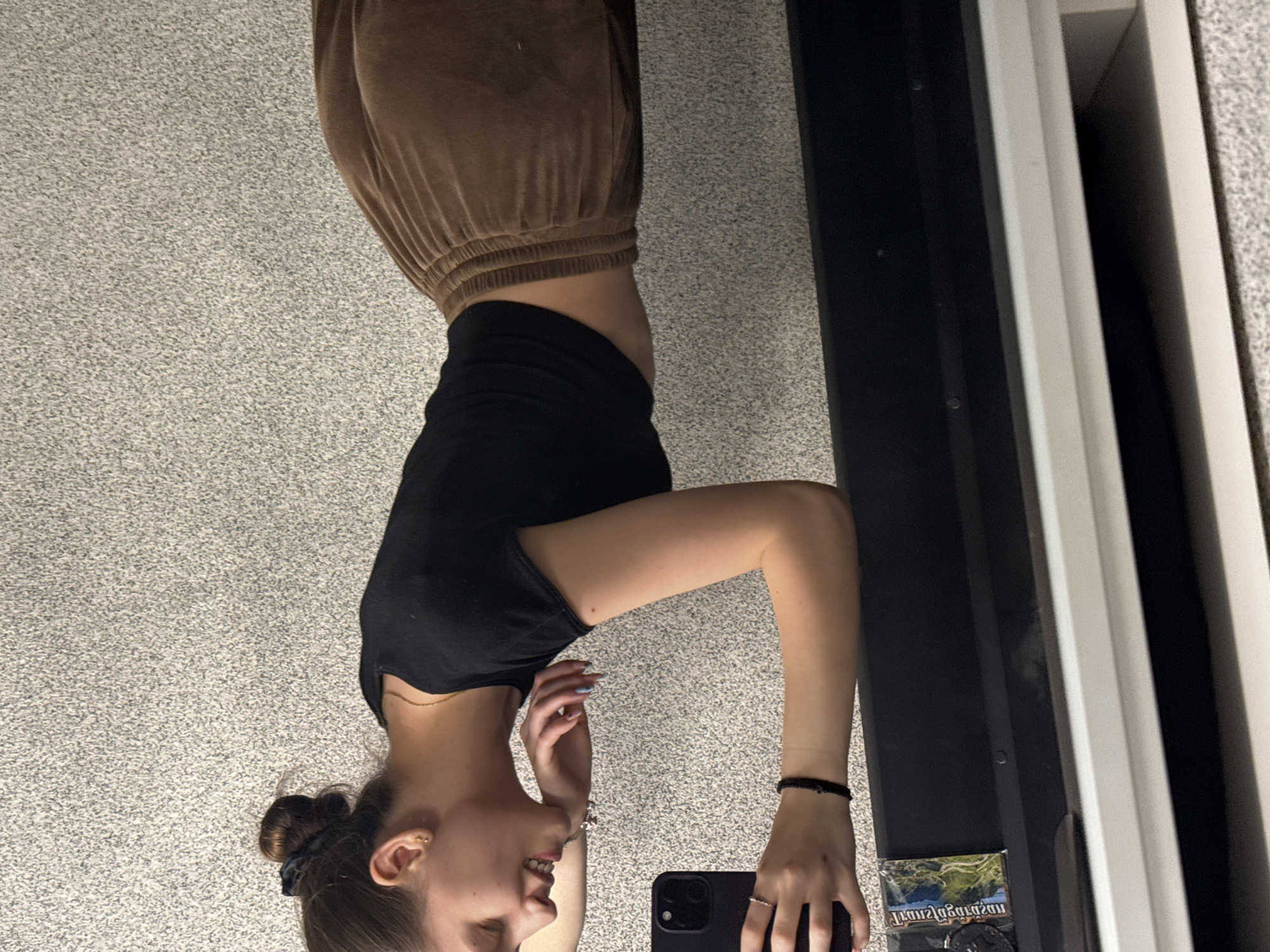 A young woman is seen in a reflection. She has brown hair pulled back in a bun and is wearing a black t-shirt and brown sweatpants. She is holding a black cell phone. The reflection is in a mirror near a wall. The location and city is not possible to determine based on the visual data of the image.