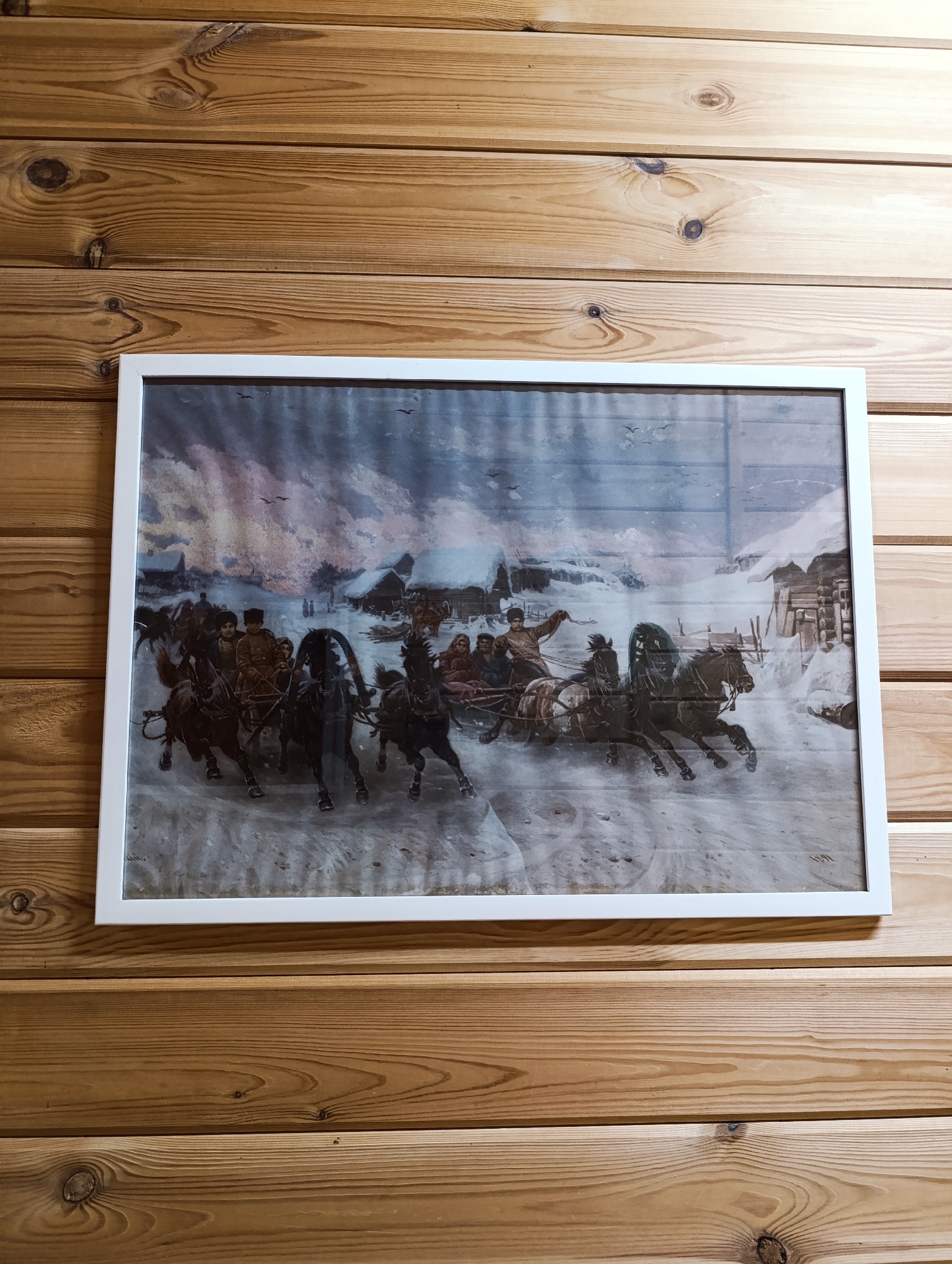 The image shows a framed print hanging on a wooden-paneled wall. The print depicts a winter scene of a horse-drawn carriage or sleigh racing through a snowy landscape. Several horses pull the carriage, and the people within are dressed in what appears to be traditional clothing. In the background, there are structures resembling houses or buildings, also covered in snow. The artwork has a distinct historical or classical style. The wall behind the print is composed of horizontal wooden planks, with visible knots and grain patterns. The location and city are not determinable from the image.