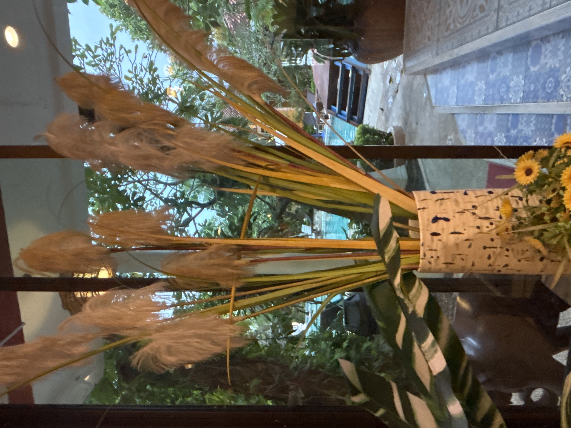 A vase with tall, dried grass plumes sits in the foreground of a room, looking out through a large glass window to a tropical outdoor scene. The grass plumes are light brown and feathery, with some appearing to be lit from behind by warm indoor lighting. The vase is cylindrical with a speckled pattern of dark blue and cream. To the left of the vase, large, dark green leaves with white stripes are visible.
Beyond the glass, a bright turquoise swimming pool is visible, with a wooden deck and blue steps leading into the water. Lush green foliage, including palm trees and other tropical plants, surrounds the pool area. The sky above is a soft blue, suggesting it is daytime. The overall impression is a tranquil resort or residential setting in Chalong, Thailand. There are no people visible in the image.