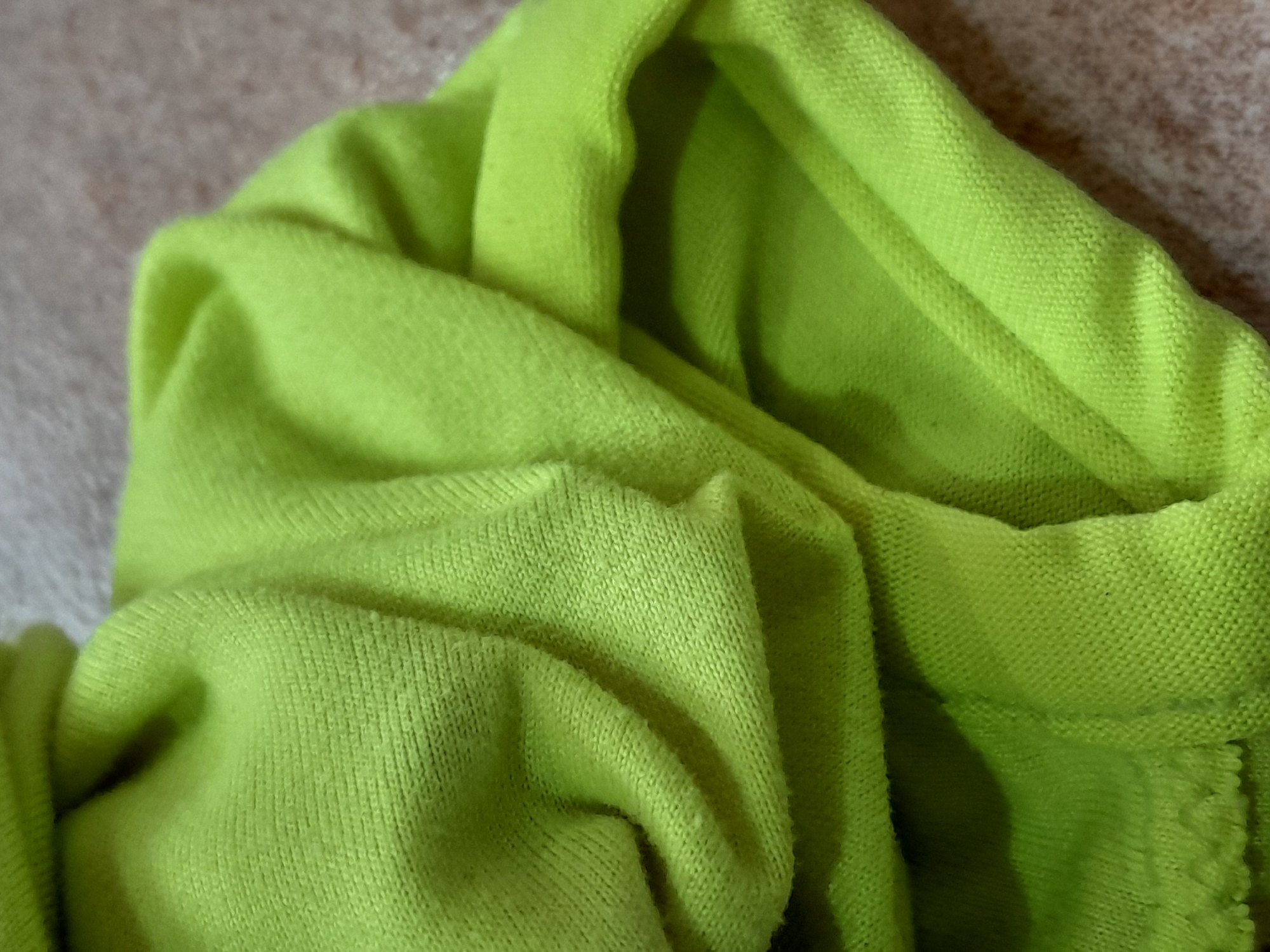 The image shows a close-up, abstract view of crumpled neon green fabric, possibly a garment like a sweatshirt or hoodie. The fabric has a textured knit surface with visible stitching and folds creating shadows and highlights. The background appears to be a textured, muted surface, possibly a carpet or upholstery. The scene is not identifiable as being in Santa Ana, Venezuela or any specific location. There are no people, actions, or interactions depicted.