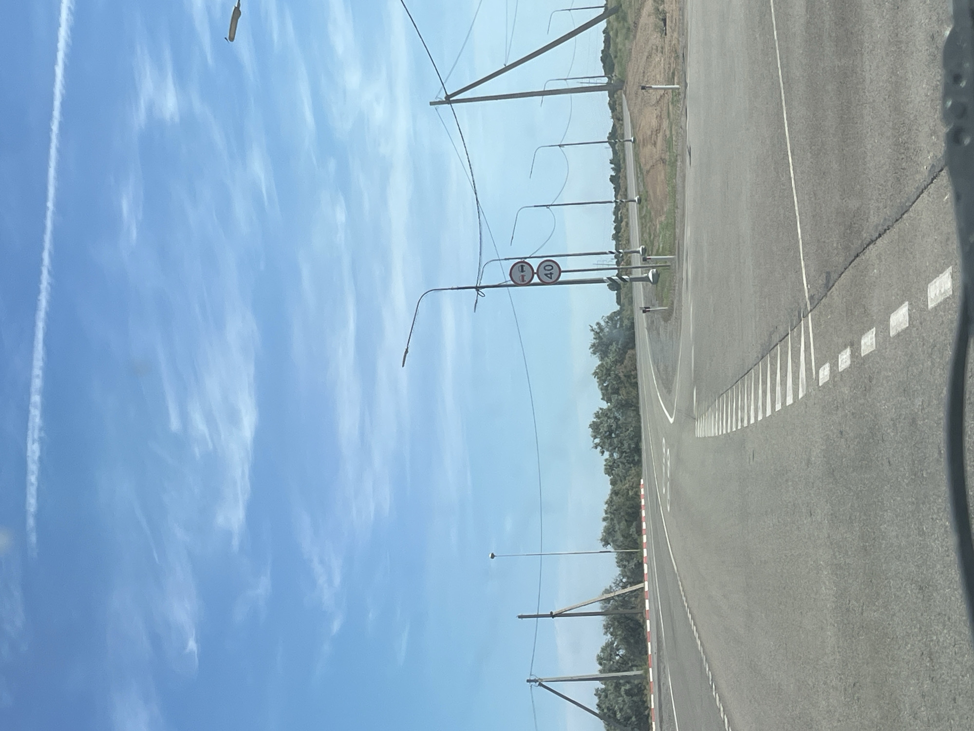 The image depicts a road in a rural or semi-rural setting. The road is paved and has white lane markings, including dashed lines and a solid white line separating lanes. On the right side of the road, there are white squares painted on the asphalt at intervals, possibly as markers or for reflecting light. The road appears to curve slightly to the right.
Along the left side of the road, there are several tall poles, likely for streetlights or power lines. Two of these poles are close enough to the road to show visible signage. One sign is a circular red and white sign with a horizontal bar, indicating a prohibition. Next to it is another circular sign with the number "40" in black on a white background, suggesting a speed limit of 40. Further down the road, more poles are visible, some with overhead wires.
The background shows a dry, dusty landscape with sparse vegetation and trees, particularly on the right side of the road. The sky is a clear, bright blue with scattered wispy white clouds, suggesting a sunny day. There are also some contrails visible in the sky. The overall impression is of a road in an open area, possibly leading to or from a less populated region. The time of day appears to be daytime, likely late morning or early afternoon due to the bright sunlight. No people are visible in the image.