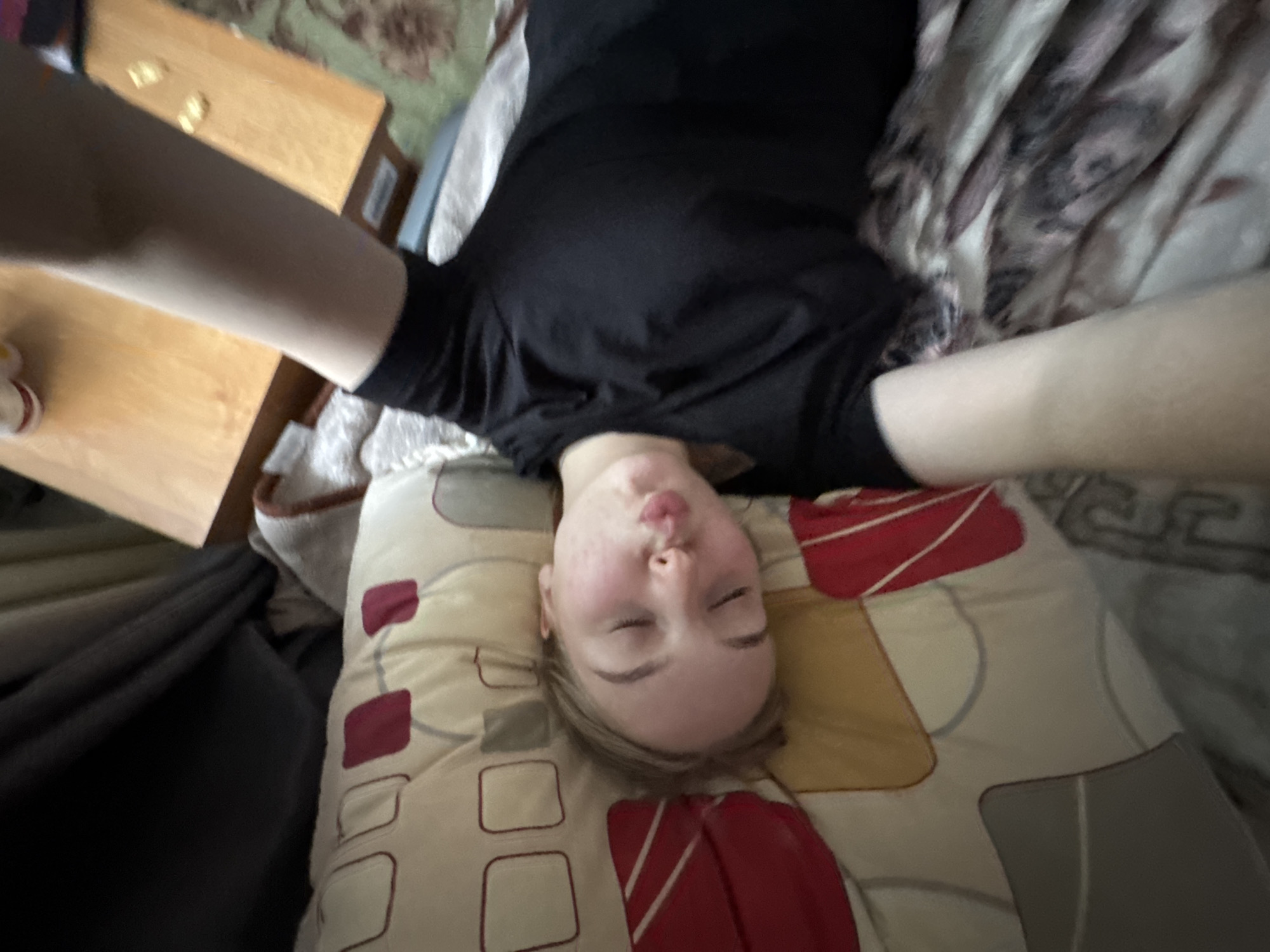 An overhead shot captures a young individual lying on their back, seemingly on a bed or sofa, in an indoor setting. The person, with fair skin and light brown or blonde hair, has their eyes closed and lips puckered into a "kissing" or "fish" face. They are wearing a black t-shirt or top, with their arms extended upwards towards the camera, indicating they are taking a selfie.
Beneath their head is a pillow featuring a geometric pattern with large squares and rectangles in shades of red, yellow, cream, and light grey. The person's body rests on a bedspread or blanket with a patterned design, possibly floral or abstract, in greenish-brown hues. To the left of their head, a wooden surface, likely a table, is visible, on which a few small, light-colored objects are scattered. Another blanket or fabric, white with dark grey or black patterns, is draped near their right arm. The overall atmosphere appears relaxed and playful. No text is visible in the image.