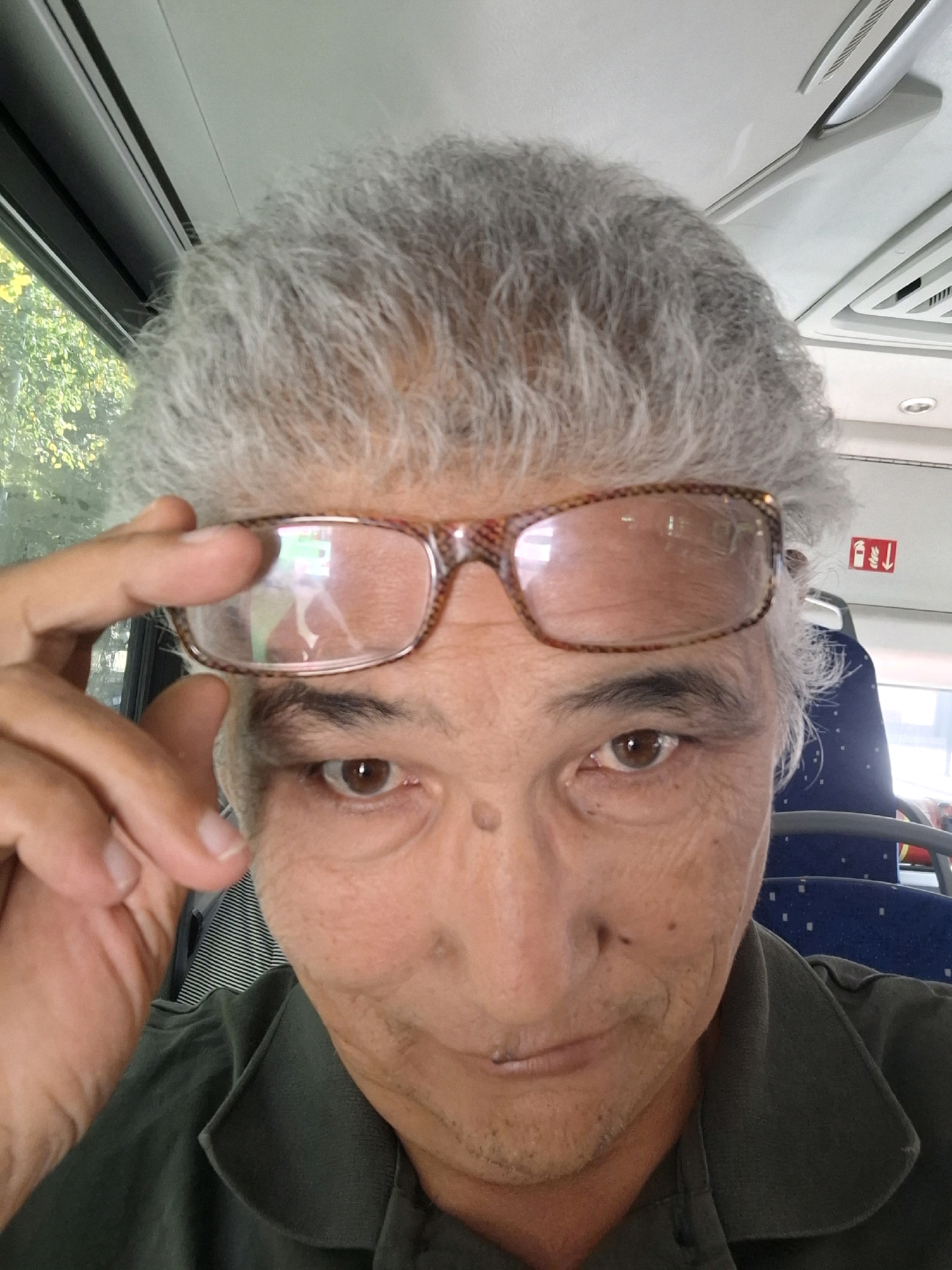 This is a close-up, selfie-style photograph of an older man with grey, curly hair, wearing glasses. He is inside a vehicle, likely a bus or train, as indicated by the presence of blue upholstered seats with white speckles visible in the background and a ceiling with ventilation grilles. The man is wearing a dark green collared shirt. He is holding his glasses up with his right hand, revealing his eye. The lighting suggests it is daytime. The image does not provide any visual cues or landmarks that would specifically place the scene in Almaty, Kazakhstan.