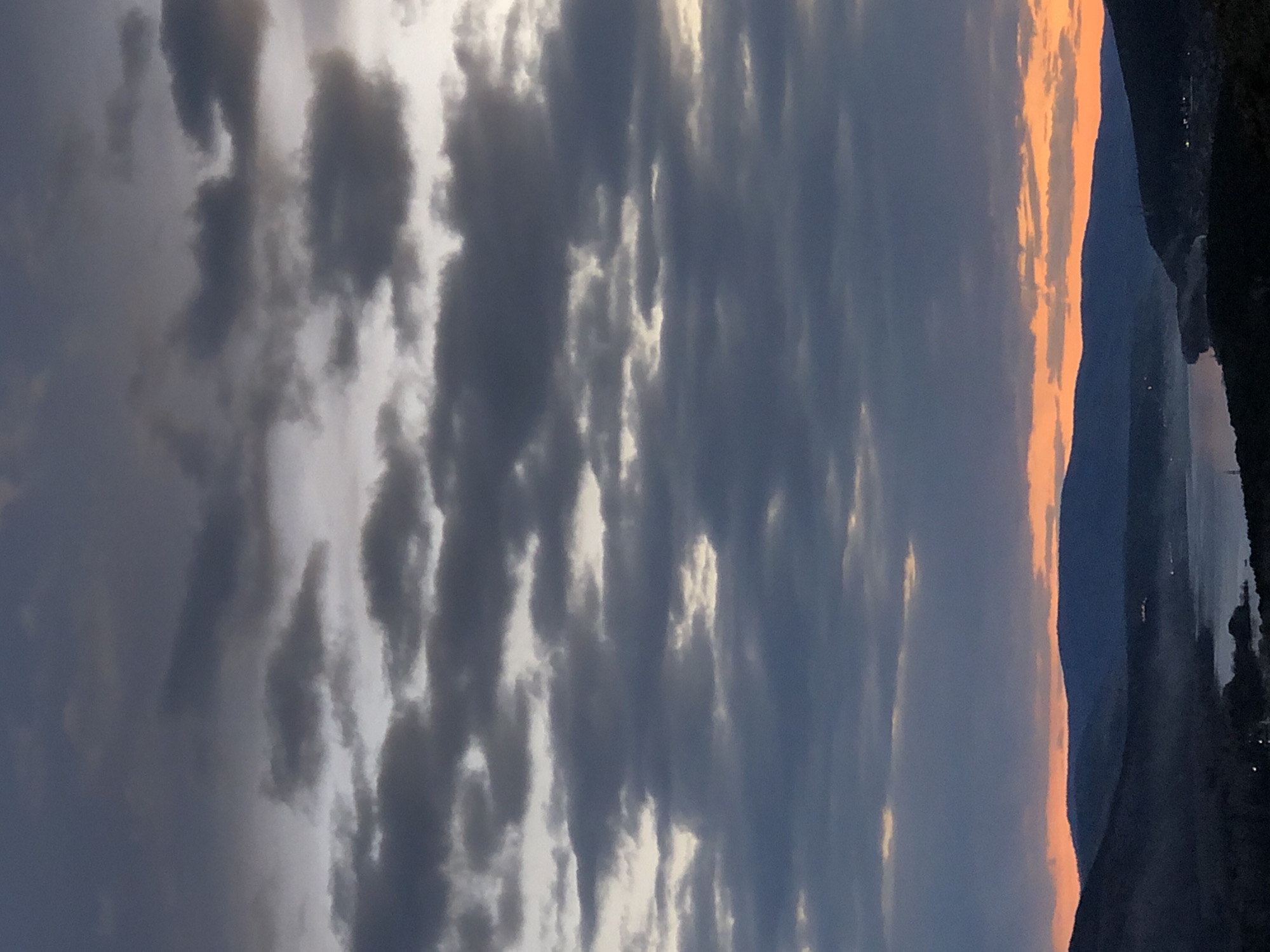 The image depicts a landscape scene at sunset or sunrise. The sky is filled with a mix of dark, gray, and white clouds. On the right side of the image, there are silhouetted mountains or hills with a strip of orange and red light along the horizon, indicating the sun's position. A body of water, possibly a river or lake, is visible in the valley between the mountains, reflecting some of the light. Based on the landscape features, the image likely captures a view along the Yenisey River near Krasnoyarsk. The image lacks people and immediate actions.
