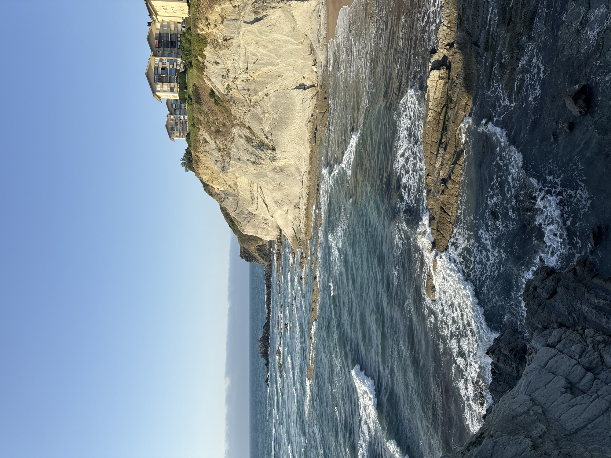 This is a scenic, eye-level shot of the coastline in Sopela, Spain, featuring a cliff face overlooking the ocean. The sky is clear and blue, with a hint of clouds on the horizon. The cliff, made of light-colored rock, is topped with several modern buildings. The ocean is a deep blue-green, with white waves crashing against the rocky shore and scattered rock formations in the water. The sun is casting a warm light on the scene, highlighting the textures of the rocks and the waves. There are no people or animals visible in the image.