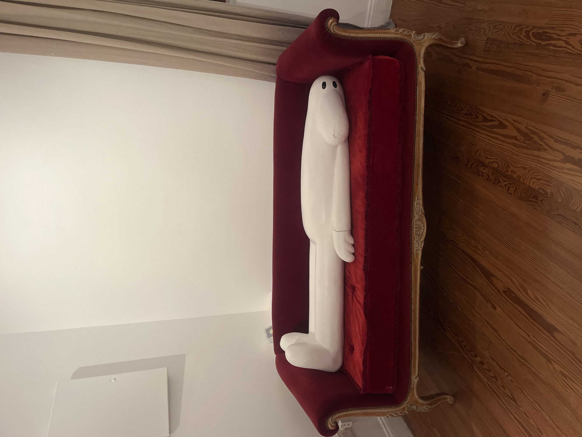 A full-length white cartoon-like sculpture rests on a red, tufted chaise lounge with gold trim in what appears to be an indoor setting. The sculpture is humanoid with a rounded head, small black eyes, arms at its sides, and large feet. The chaise lounge is positioned at an angle against a white wall, with a light-colored curtain visible at the top of the frame. Wood flooring is present to the right of the chaise lounge. The specific location in the city of Buenos Aires, Argentina, cannot be determined from this image.