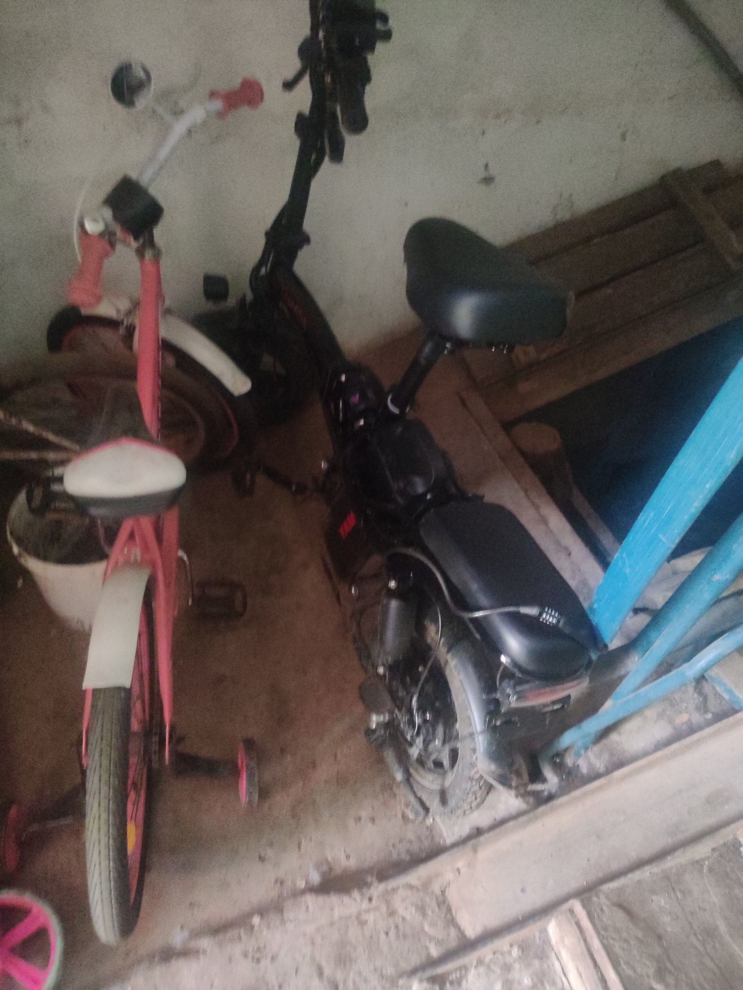 The image shows a pink child's bicycle and a black electric scooter parked indoors. The bicycle is positioned on the left side of the frame, with its front wheel visible. The electric scooter is on the right, and its seat and rear wheel are clearly discernible. The environment appears to be a garage or storage area, with concrete flooring and wooden elements visible in the background. There are no people present in the image. The lighting is dim, suggesting an indoor, possibly unlit space.
