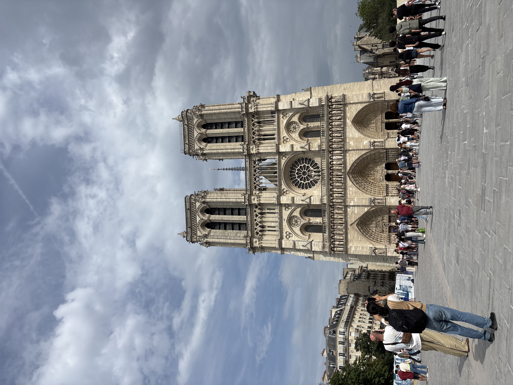 The image depicts the exterior of Notre Dame Cathedral in Paris, France, on a cloudy day. The facade of the cathedral, a magnificent Gothic structure, is the central focus, with its two prominent bell towers and intricate architectural details, including sculptures and arched doorways. A large rose window is visible on the upper level of the facade.
In the foreground, a paved plaza is populated by numerous tourists and locals. Several individuals are engaged in various activities, such as taking photographs of the cathedral, walking, and conversing. On the left side of the image, two people are standing close together, with one person holding up a phone, seemingly taking a picture. The ground is paved with large, rectangular stones.
In the background, to the left of the cathedral, there are several Parisian buildings with mansard roofs, characteristic of the city's architecture. The sky is filled with a mix of light and dark clouds, suggesting a dynamic weather pattern. The overall scene captures a moment of public life and tourism in front of a globally recognized historical landmark.