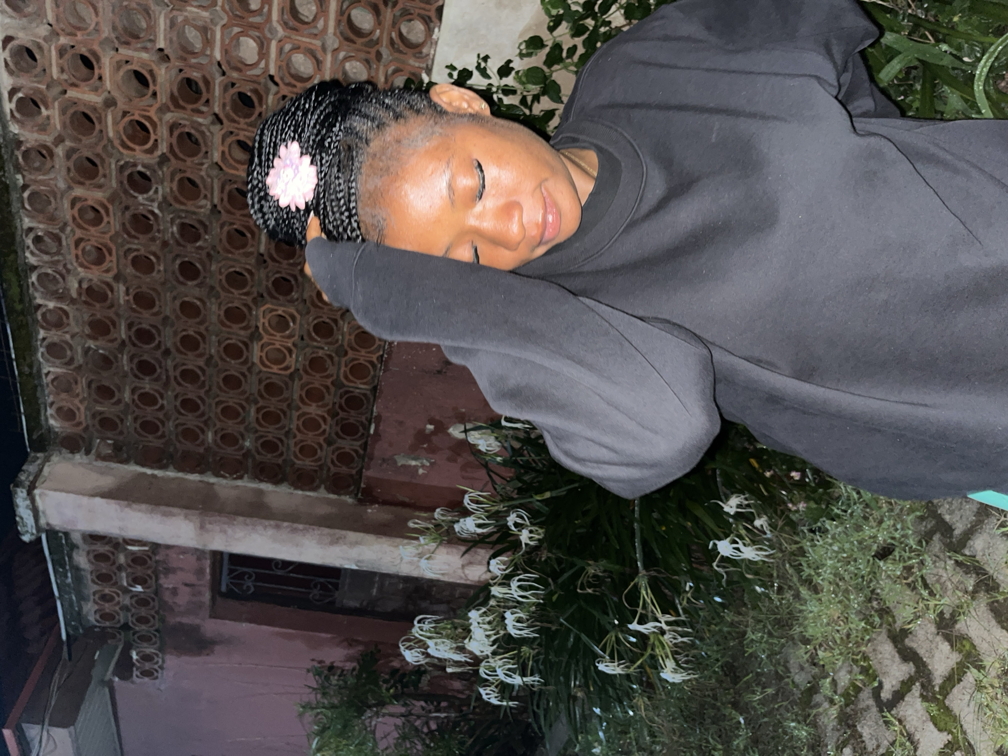 A young Black woman with braided hair adorned with a pink flower accessory is captured in a medium shot outdoors at night. She is wearing a dark gray long-sleeved sweatshirt and has her right arm raised, touching her head. Her eyes are closed, and her head is tilted down, suggesting a moment of reflection or rest. The background features a patterned brick wall with circular openings, a pink-painted wall, and a cluster of white spider lilies in full bloom. The ground is paved with interlocking gray bricks, some of which are wet and reflect light, indicating recent rain. Green foliage surrounds the flowers and extends into the lower right portion of the frame. The lighting is dim, consistent with nighttime. The scene appears to be within a residential or garden area.