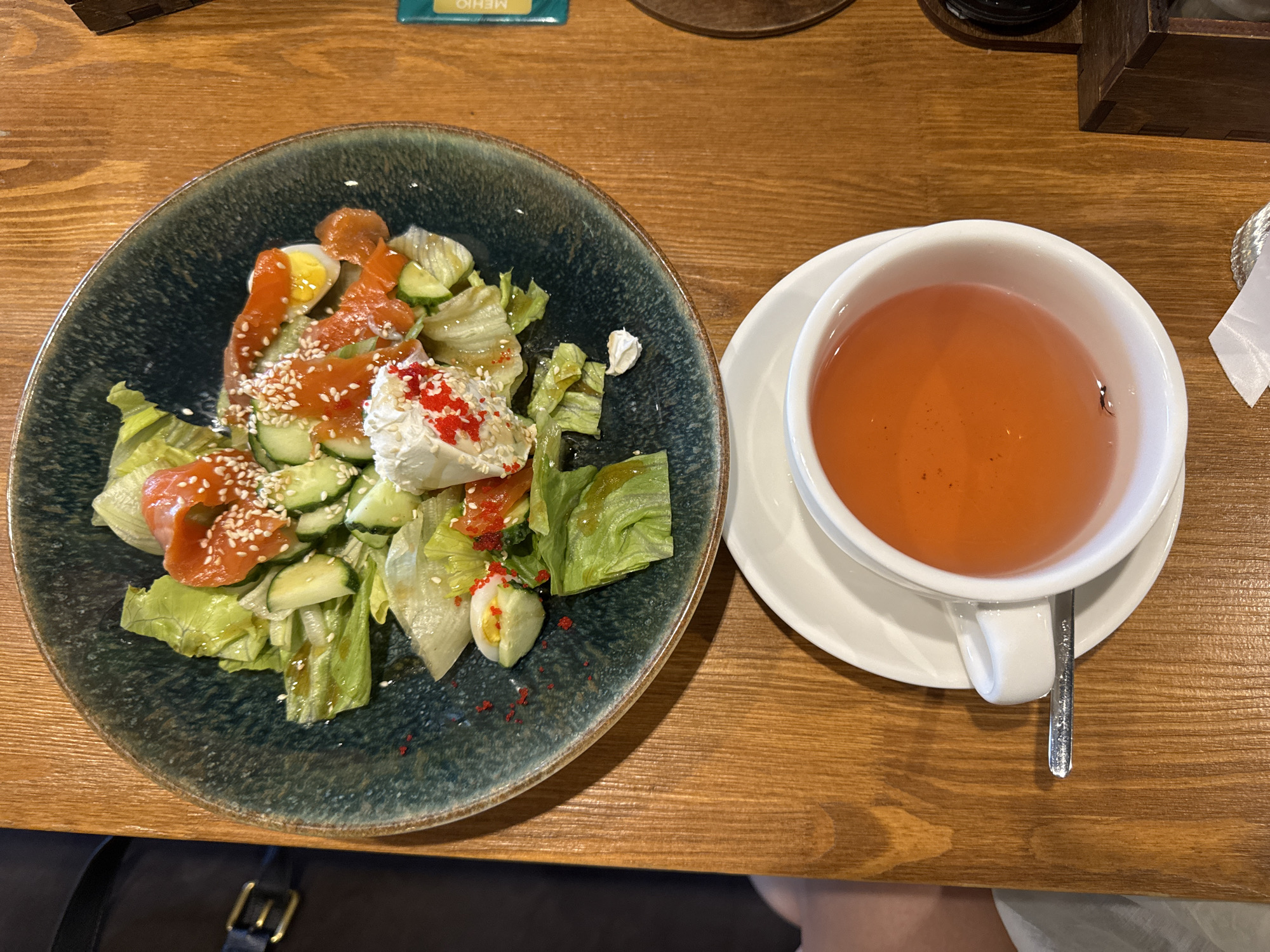 The image shows a close-up, high-angle view of a table setting on a wooden surface. On the left is a dark, shallow bowl containing a salad. The salad appears to be made of lettuce, sliced cucumbers, quail eggs, and pieces of smoked salmon. It is garnished with sesame seeds and small red roe or beads. On the right is a white teacup with a saucer, filled with a reddish-orange liquid, likely tea. A spoon rests on the saucer, partially submerged in the tea. In the background, a corner of a wooden box and a piece of paper with Cyrillic text are visible. The overall scene suggests a meal or refreshment break in Kazan, Russia, possibly at a cafe or restaurant. No people are explicitly visible, though a knee and foot are faintly discernible at the bottom right, indicating a person is present and seated at the table. The lighting is natural and diffused, casting soft shadows.