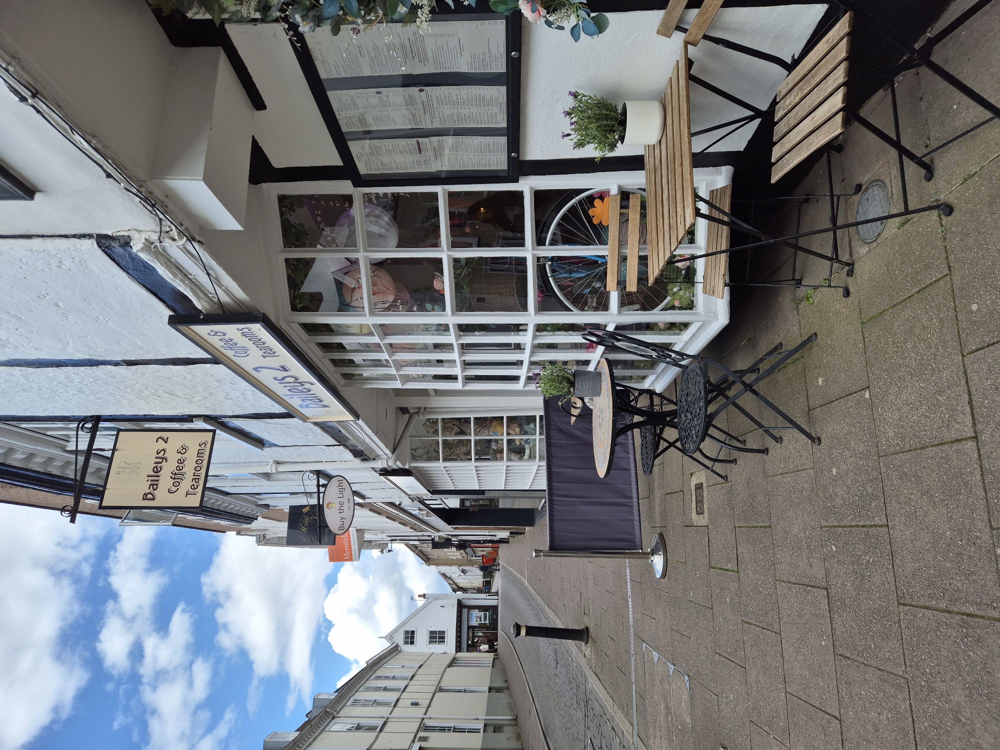 The image captures a street scene in West Suffolk, United Kingdom. Buildings line both sides of the street, featuring various architectural styles, including white-painted facades and traditional timber framing. Signage for "Baileys 2 Coffee & Tearooms" and "Buy the Light" can be seen. Outdoor seating is arranged outside a building, with chairs and tables set up on the paved sidewalk. A crowd control barrier is positioned on the right side of the image. Overhead, a blue sky with scattered clouds is visible.