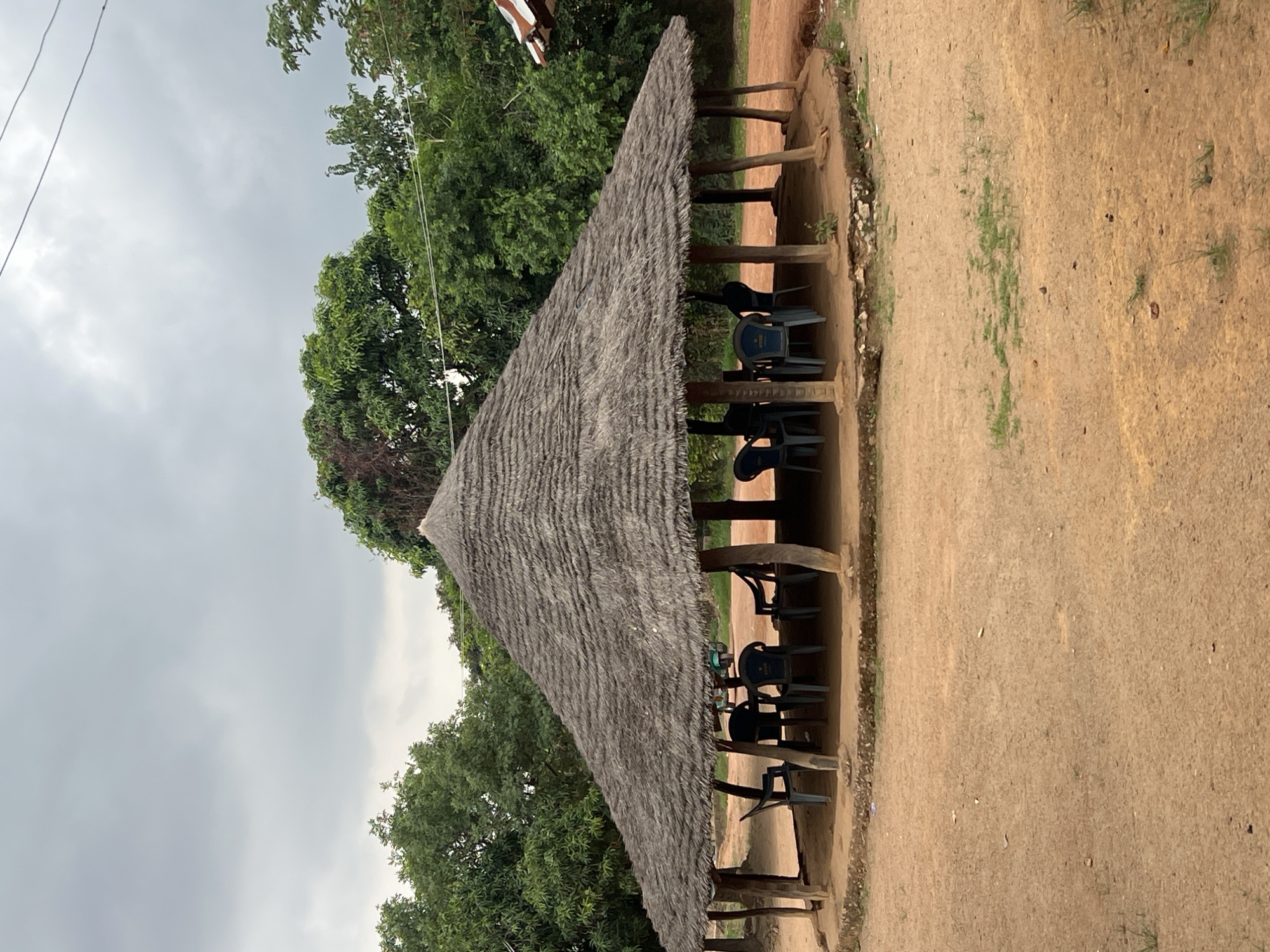 The image displays an outdoor scene featuring an open-sided shelter with a traditional thatched roof, set in a rustic environment under an overcast sky. The photograph is oriented vertically, showing the ground to the right and the sky and trees to the left when viewed in its correct horizontal orientation.
The central subject is a substantial shelter with a sharply angled, conical-like thatched roof made of dried straw or grass, giving it a textured, grey-brown appearance. The roof is supported by numerous dark wooden posts, creating an inviting, shaded area beneath. Inside the shelter, a series of dark blue and black plastic chairs are arranged around low wooden benches or tables, suggesting a casual dining or gathering spot. On the backrests of some blue chairs, a distinctive white oval logo with a star symbol and the word "STAR" is visible, indicating a branded beverage.
The ground surrounding the shelter is unpaved, consisting of reddish-brown dirt or packed earth, with scattered small patches of green grass. Beyond the shelter, lush green trees with dense foliage form a natural backdrop, emphasizing the outdoor setting. The sky above is entirely covered with grey clouds, indicating an overcast or cloudy day.
No people are present in the image, but the setup of chairs and tables implies a space designed for social interaction, rest, or refreshment. The scene evokes a sense of calm and simplicity, typical of local establishments in many parts of Nigeria, aligning with the provided location context of Jos. The lighting suggests it is daytime, though the lack of direct sunlight points to a non-sunny weather condition.