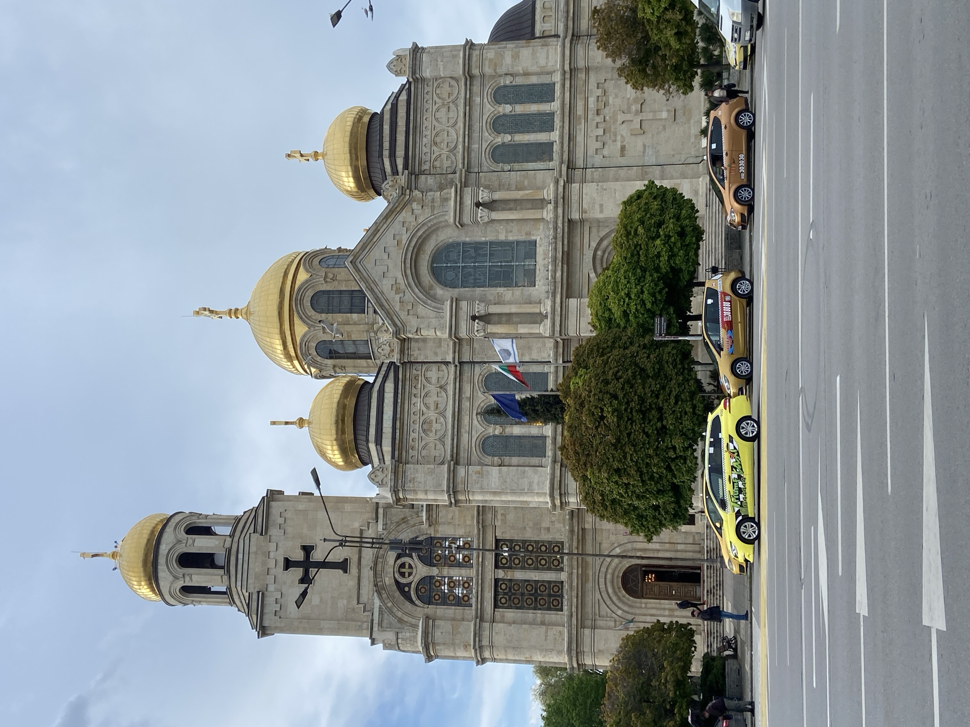 The image shows the Dormition of the Theotokos Cathedral in Varna, Bulgaria. The cathedral is a large stone building with multiple golden domes and crosses on top. Flags are displayed on the exterior. There are three taxis parked along the street in front of the cathedral. A few people are present near the entrance of the building.