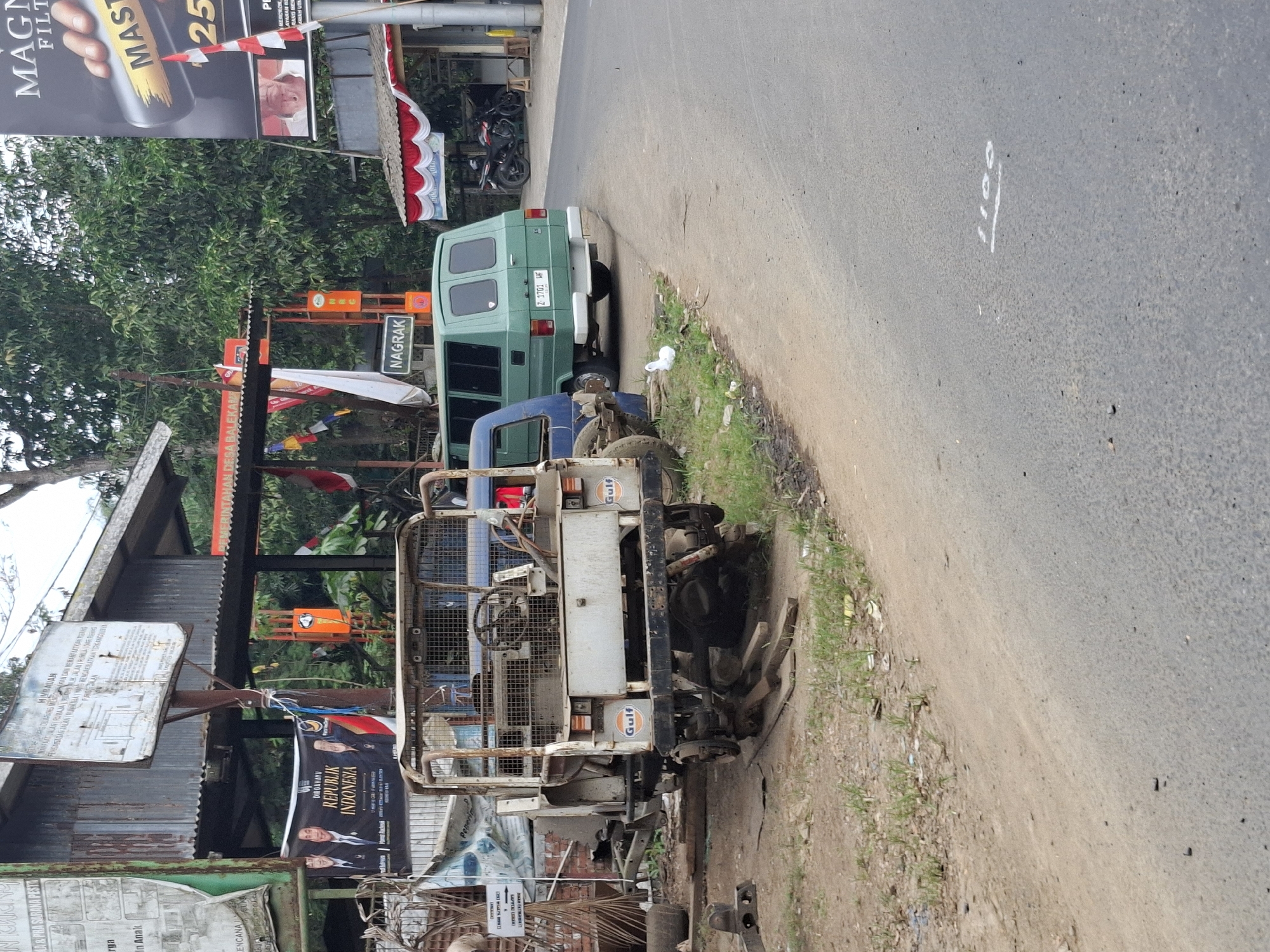 The image depicts a roadside scene in Balekambang, Indonesia. In the foreground, a rusted, white utility vehicle, possibly a jeep or similar utility vehicle, is parked on a dirt and gravel area next to a paved road. The vehicle appears to be in disrepair, with exposed metal and missing parts. Next to it, a green van is parked on the paved road. The van has tinted windows and appears to be in good condition. Further back, a makeshift structure with a corrugated metal roof is visible, adorned with various banners and flags, including a black banner with the text "DIRGAHAYU REPUBLIK INDONESIA." Trees and foliage line the background. To the right of the scene, a large billboard advertising "MAGNA FILTER" is visible. The road has some white markings, including the number "1100" painted on the asphalt. The overall impression is that of a somewhat neglected roadside area, possibly a business or a workshop, located in a tropical or subtropical environment. No people are clearly visible in the image.