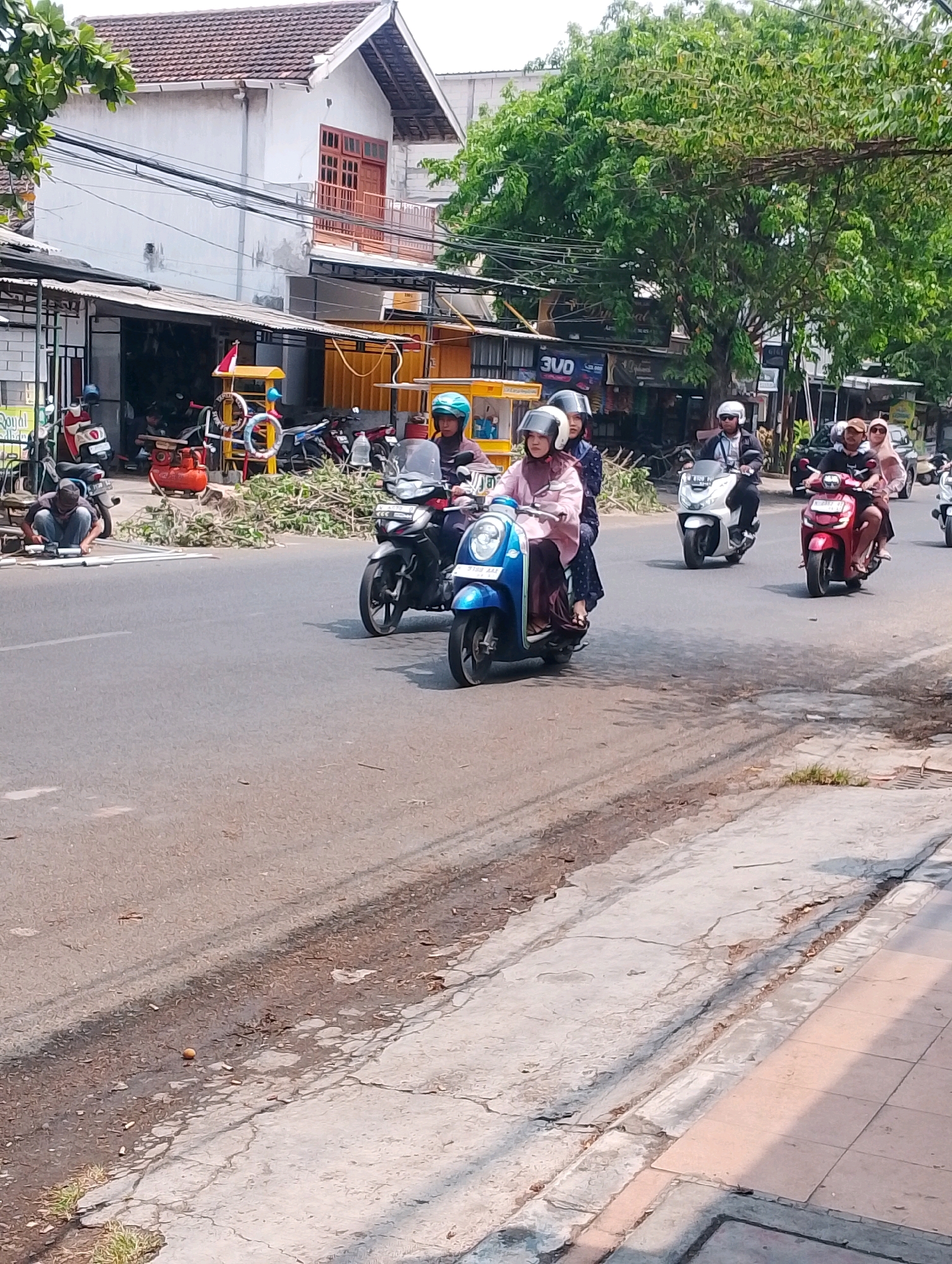 In Probolinggo, Indonesia, on a sunny day, a street scene unfolds with multiple individuals riding motorcycles. In the foreground, a blue scooter carries two people, a woman in a pink hijab and a passenger behind her. To their right, a red scooter is ridden by a man wearing a light-colored hijab, with a woman in a pink hijab as the passenger. Further down the road, a white scooter is being ridden by a man in a dark jacket and helmet, and behind him, another motorcycle carries a couple. The street is lined with buildings and trees, and there are various shops visible, including one with yellow awnings. A man is kneeling on the side of the road, appearing to be working on something. A pile of branches lies on the side of the street near a red gas tank. The asphalt road shows signs of wear, with some cracks and debris. A concrete sidewalk runs along the right side of the road, with some areas appearing damaged.