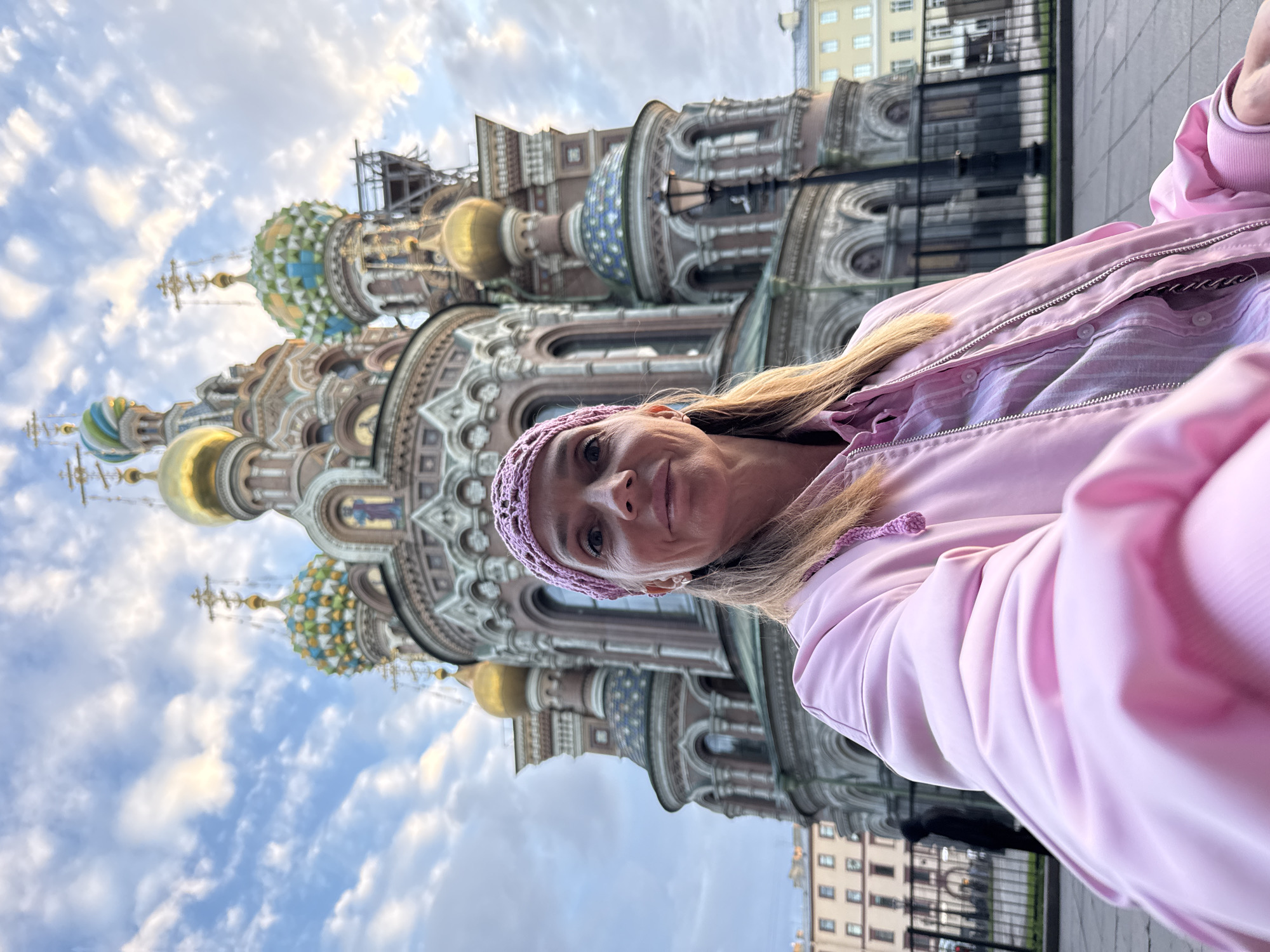A woman, wearing a pink knitted headband and a pink jacket over a pink striped shirt, takes a selfie in front of the Church of the Savior on Spilled Blood in Saint Petersburg, Russia. The church, with its ornate onion domes and intricate architecture, is the dominant background element. The sky above is partly cloudy with hints of sunset. In the background to the left, a silhouette of a person in dark clothing is visible behind a black metal fence. Other buildings in a lighter color are also visible in the distance on the right.