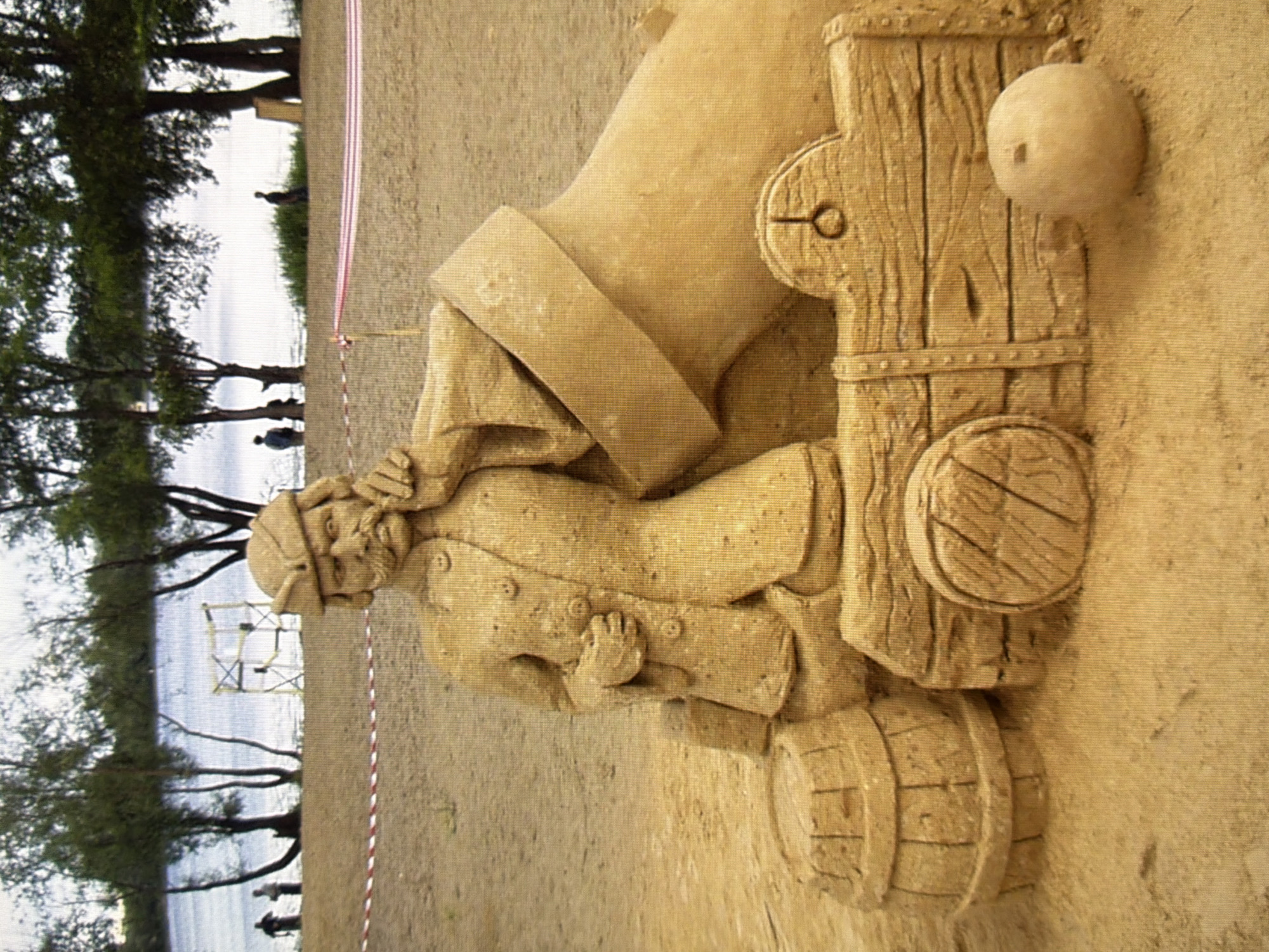 A sand sculpture of a historical figure, likely a soldier or sailor, sits on a sandy beach. The figure is depicted wearing a uniform with a hat and has a prominent mustache. He is leaning on a large mortar, which is part of a cannon mechanism. To the left of the figure are two wooden barrels, and to the right is a cannonball. The sculpture is set against a backdrop of trees and a body of water, possibly a river or lake. In the distance, there are a few people visible on the shore and a yellow construction scaffold. The scene suggests an outdoor event or festival, possibly related to historical reenactment or art exhibition. The location is specified as Kyiv, Ukraine.