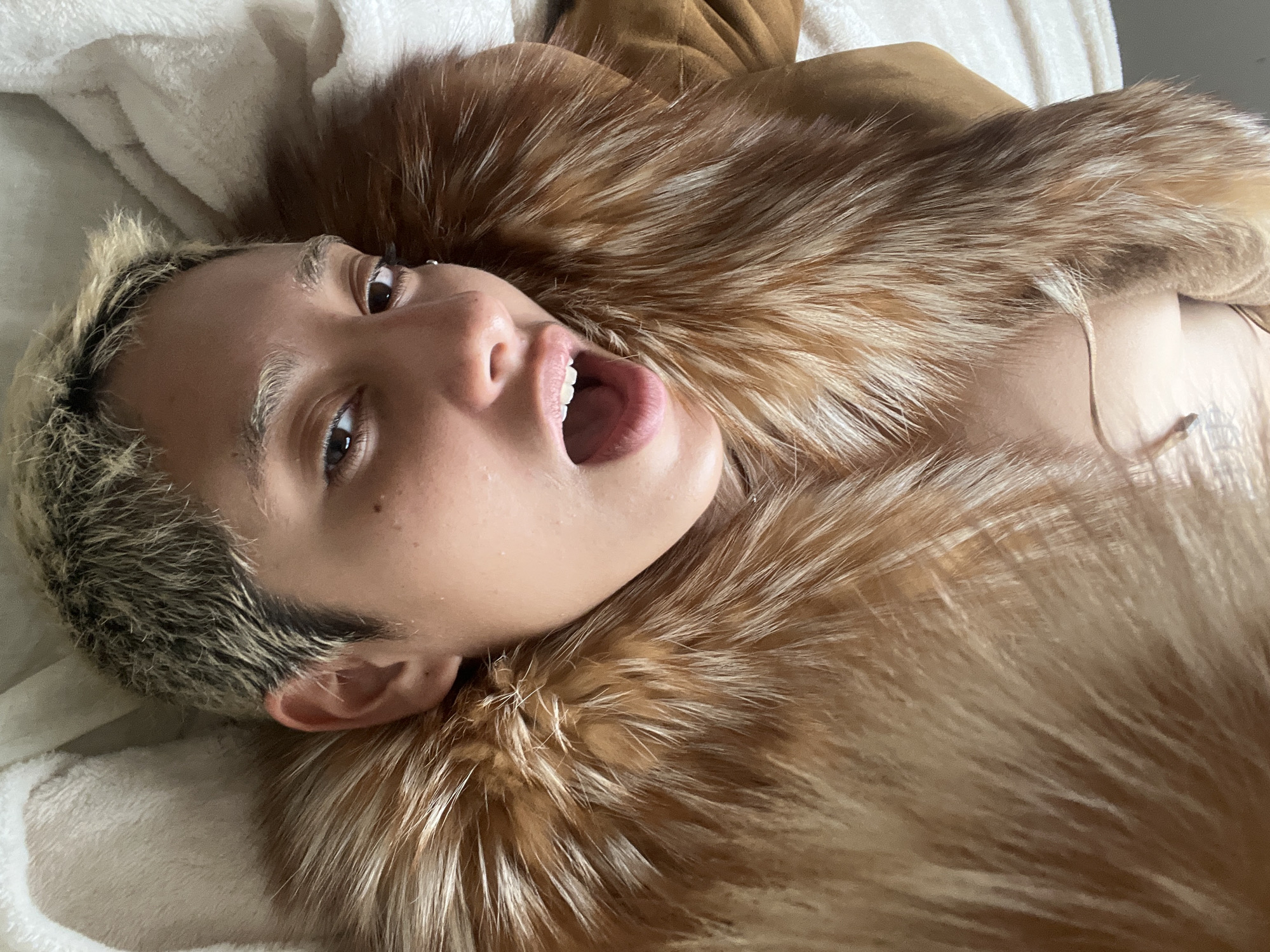 A person with short, dyed blonde hair lies on a white blanket with a tan fur collar around their neck. The person is lying on their back with their head tilted upwards and their mouth open. The person's skin tone appears to be light brown. The location is unknown.