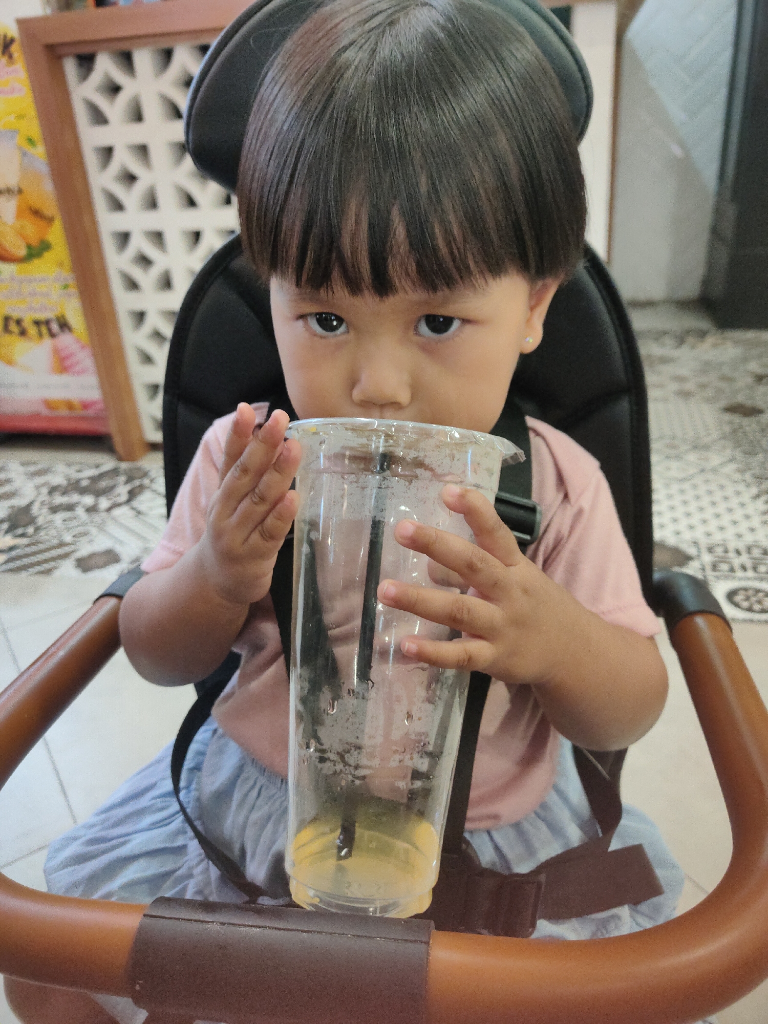 In the city of Kediri, Indonesia, a young child is seated in a high chair, holding a clear plastic cup with a straw and drinking its contents. The child, wearing a pink t-shirt and a light blue skirt, is looking directly at the camera with a neutral expression. The background shows a patterned wall and a table with a sign. The high chair has brown armrests and a black seat.