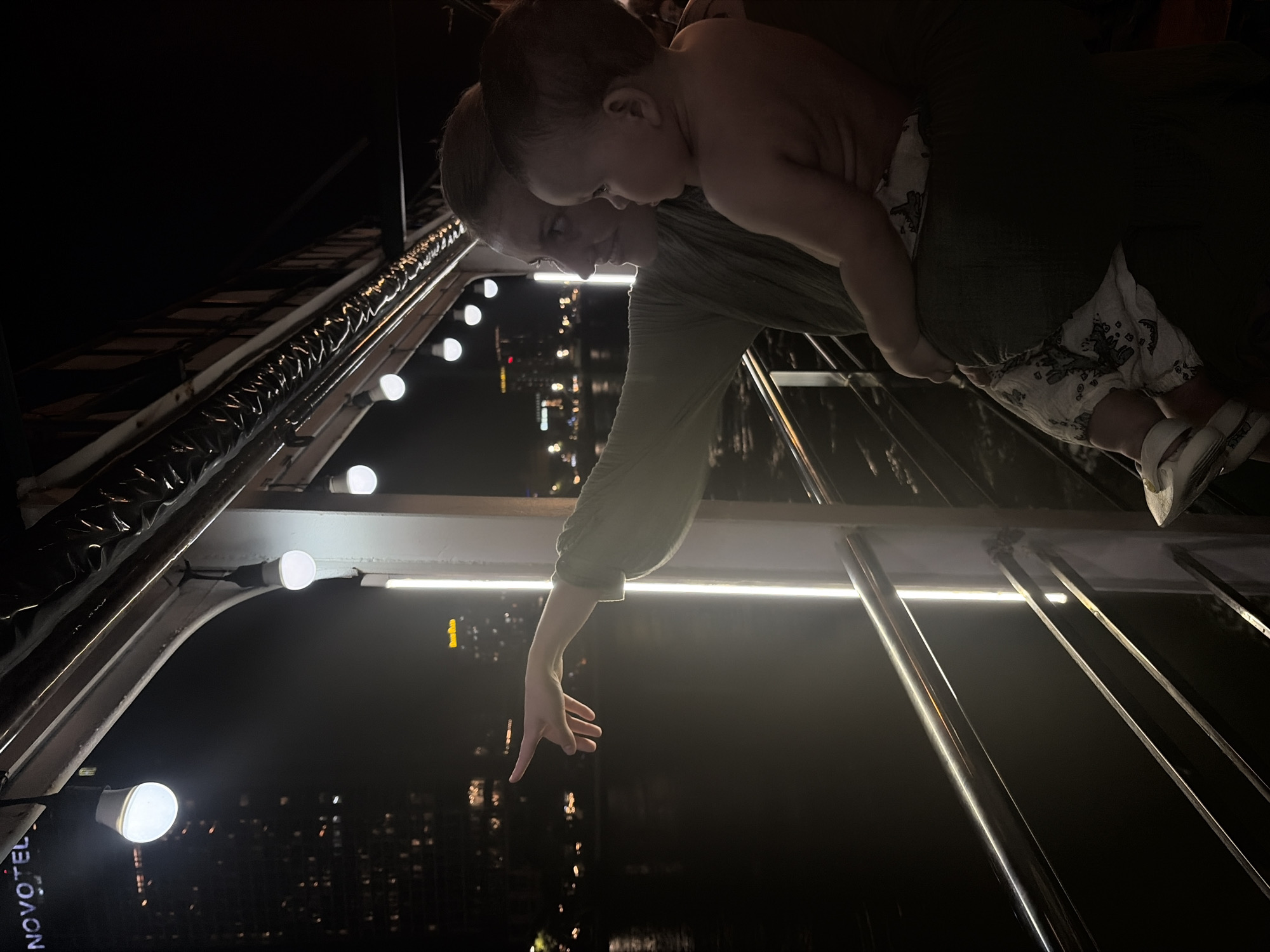The image captures an adult and a baby on what appears to be a boat or an elevated viewing platform at night, overlooking a city skyline. The adult, positioned to the right, holds the baby in front of them, both looking downwards and to the left. The adult is seen with a subtle smile, pointing with their right hand, seemingly drawing the baby's attention to something below or in the distance. The baby appears engaged, looking in the direction the adult is pointing.
The setting is dark, indicating nighttime, with the background filled with the sparkling lights of a city. The foreground features prominent metal railings and bright artificial lights – both round bulb-like fixtures and linear LED strips – illuminating the subjects and the immediate structure. The perspective suggests they are high above the water or ground, offering a panoramic view of the urban landscape.
A key visible detail is the illuminated "NOVOTEL" sign on a building in the distant city, which, combined with the provided context, places the scene in Đà Nẵng, Vietnam. This suggests the photo was taken from a position offering views of the Han River and its surrounding cityscape, possibly from a river cruise or a tall building's observation deck. The overall mood conveys a moment of shared discovery and wonder between the adult and child.