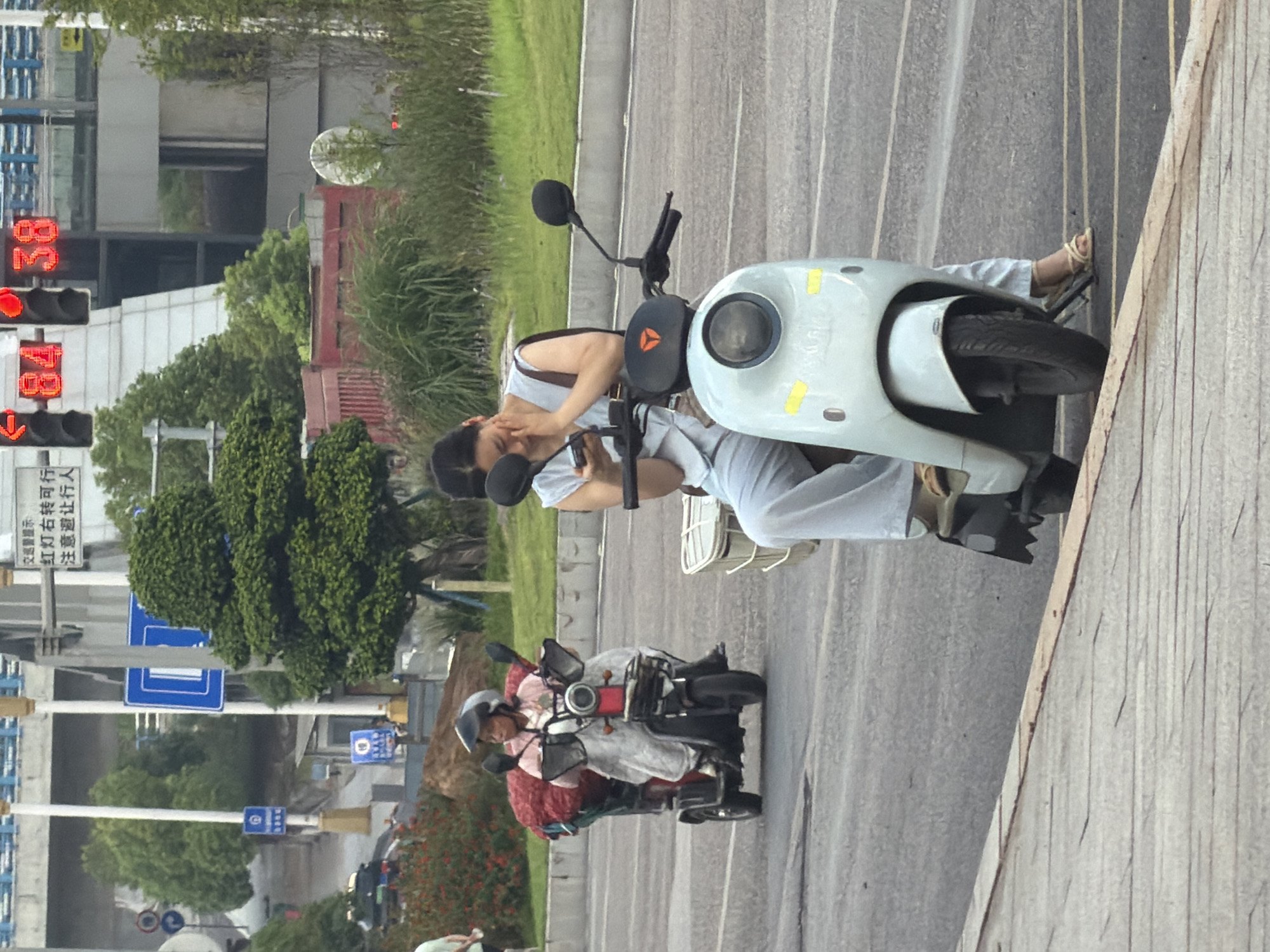 In Jiangjin, China, a woman in a light blue sleeveless top and matching pants rides a light blue electric scooter. She wears sandals and a brown bag slung over her shoulder, and she is looking at her phone while resting her head on her hand. The scooter has a white basket attached to the back and yellow reflective tape on the front. The woman is on a paved road with a white line marking the lane. Behind her, another person on a black electric scooter, wearing a helmet and a pink shirt, is also on the road, with a large red bundle strapped to the back of their scooter. The environment includes green grass, trees, and buildings in the background. Traffic lights are visible in the distance.
