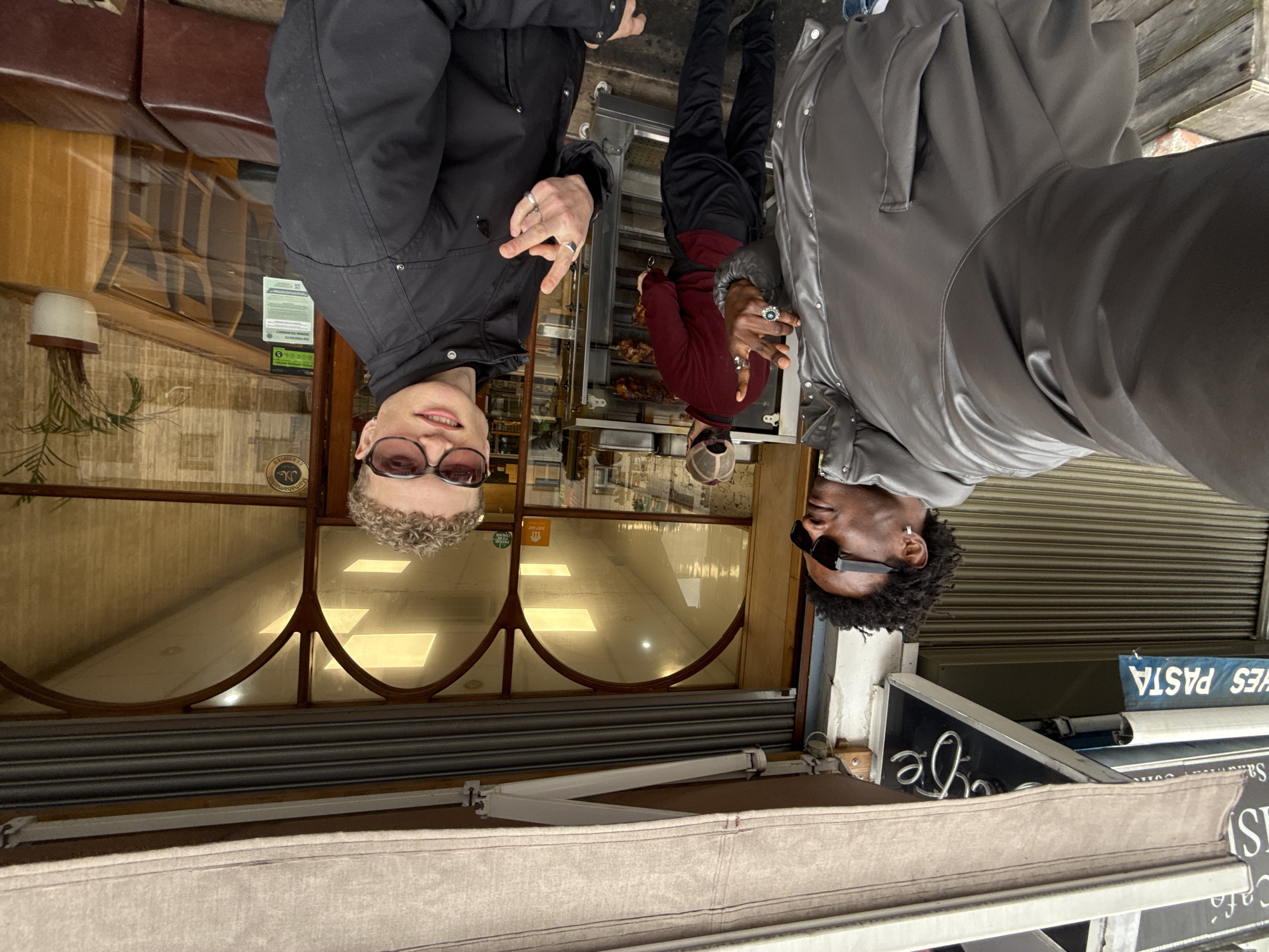 The image shows two people posing outside a restaurant in London, United Kingdom. One person is on the left side of the frame, wearing a black jacket and glasses and displaying the peace sign. To the right, the second individual wears a grey jacket, sunglasses, and also makes the peace sign. Behind them, through the restaurant's window, a person in a maroon shirt is visible tending to a rotisserie. Signs outside the restaurant advertise "PASTA" and "Cafe". The restaurant's awning is visible in the foreground.