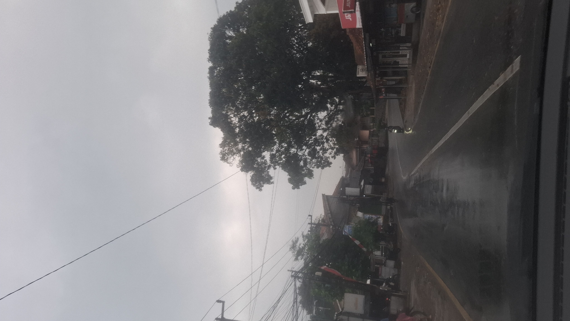 The image depicts a street scene in Cilaku, Indonesia, under an overcast sky. The road is wet, suggesting recent or ongoing rain. On the left side of the road, there are several utility poles with numerous wires crisscrossing above. Adjacent to the poles are some buildings and trees. A person, partially visible, is walking on the left side of the road, and several motorcycles are parked or being ridden in the area. On the right side of the road, there are more buildings, some with awnings, and a large tree dominates the middle ground. A motorcycle is visible on the right side of the road, with its headlight illuminated. The scene appears to be a typical street in a residential or commercial area.