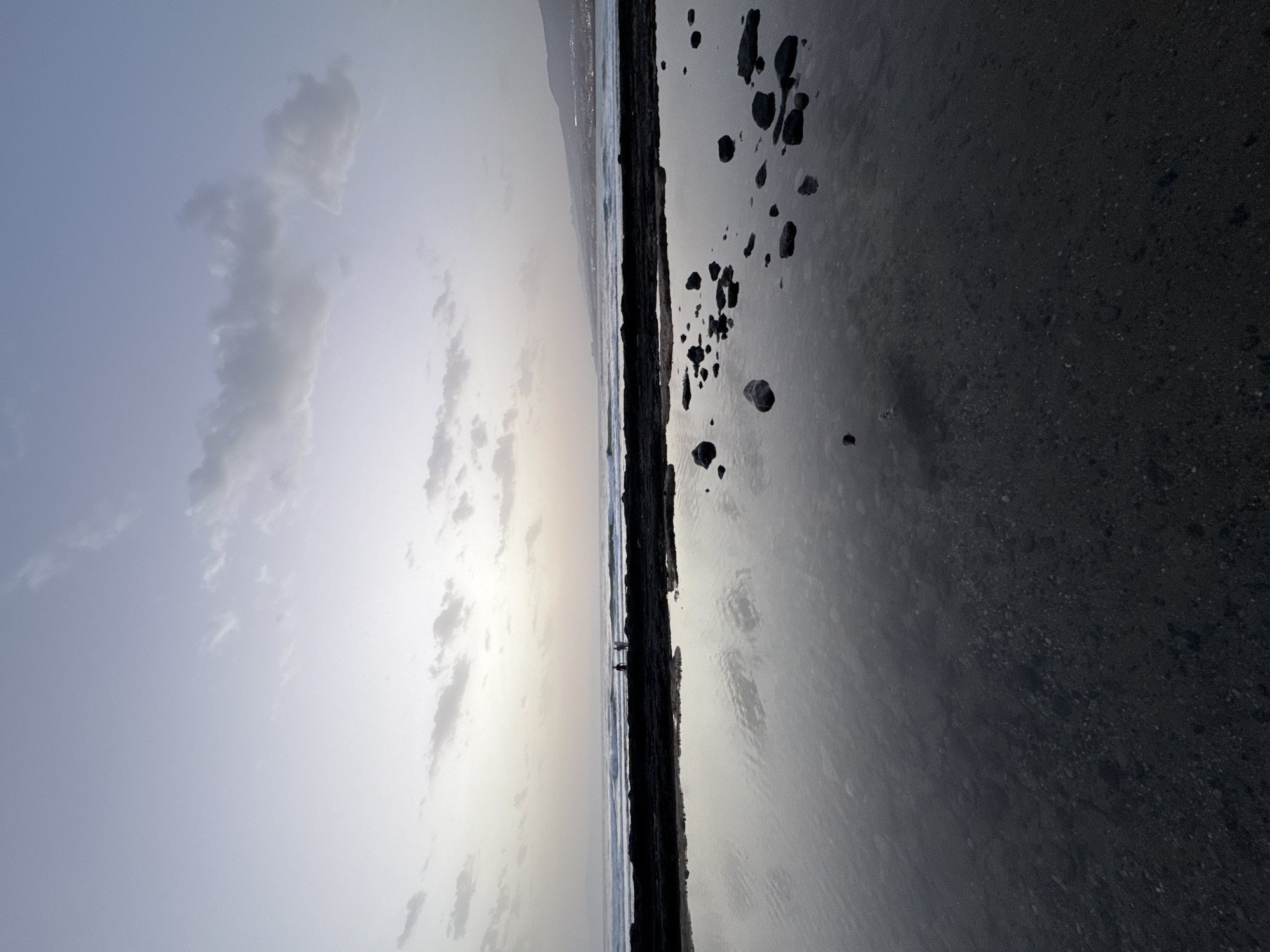 This is a vertical, wide-angle photograph taken at dusk on the coast of Arona, Spain. The foreground features a shallow tide pool filled with calm water, reflecting the sky and surrounding rocks. Several dark, smooth rocks are visible submerged in the water and scattered on the edge of the pool. A dark, rocky coastline separates the tide pool from the ocean. Gentle waves are breaking on the shore, and in the distance, two figures, one holding a surfboard, stand on the coastline. Further in the background, a distant city lights up the hazy horizon, with mountains visible beyond it. The sky is a gradient of pale blue and white, with soft, scattered clouds.