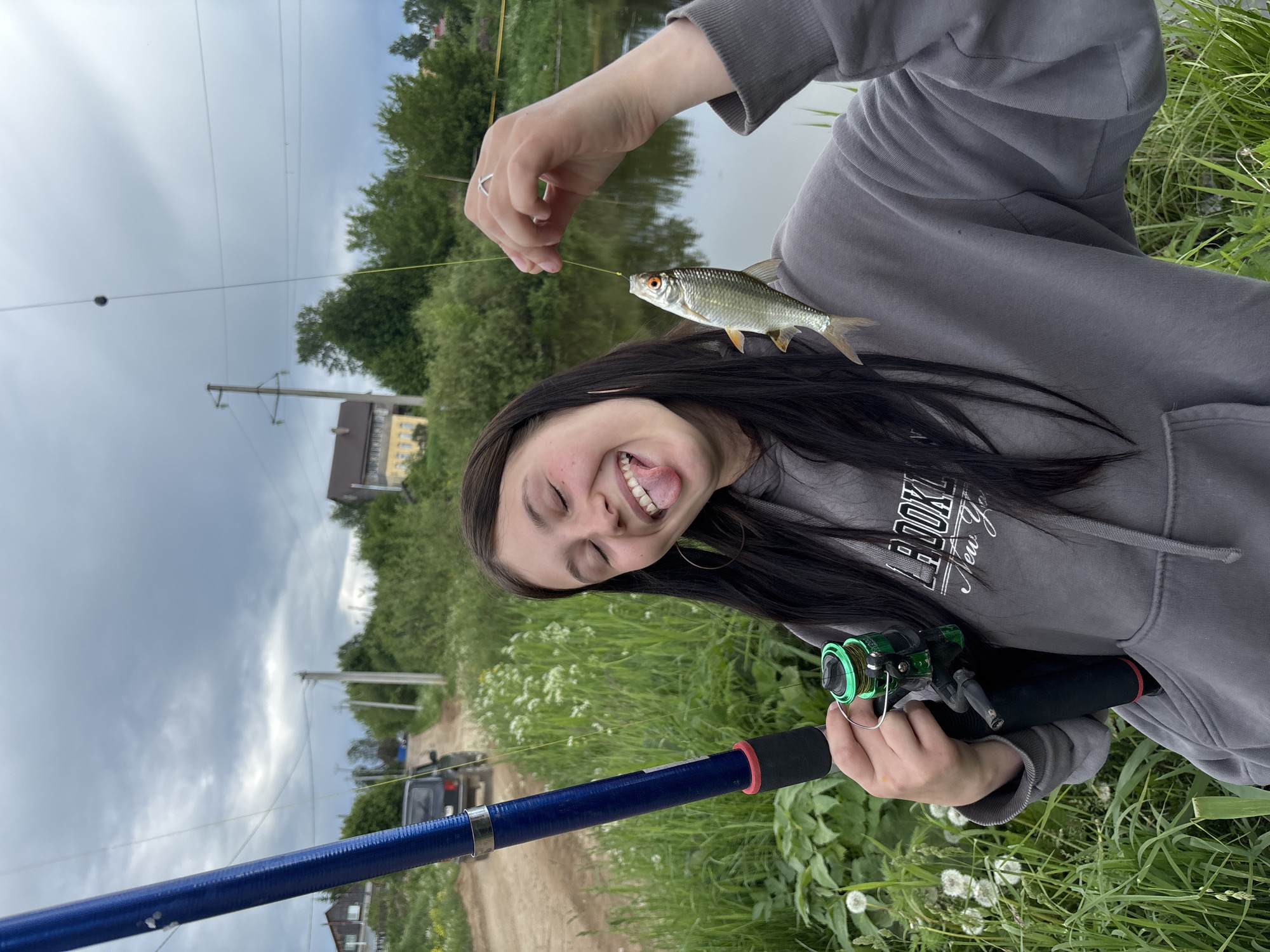 A woman with long, dark hair is holding a fishing rod with a blue shaft and green reel. She is also holding a small fish on the line near her face. She is wearing a gray hooded sweatshirt with a printed design on the chest. She has her tongue sticking out and her eyes are closed, indicating a playful or celebratory expression.
In the background, there is a body of water (likely a pond or lake), vegetation, and trees. Power lines are visible in the distance.
The location is Фоки, Russia, suggested by the presence of houses with architectural styles common in rural Russian areas and by the general type of landscape visible in the image.
A vehicle is partially visible in the background, and the scene appears to be near a dirt road. The overall impression is of a rural or semi-rural setting.