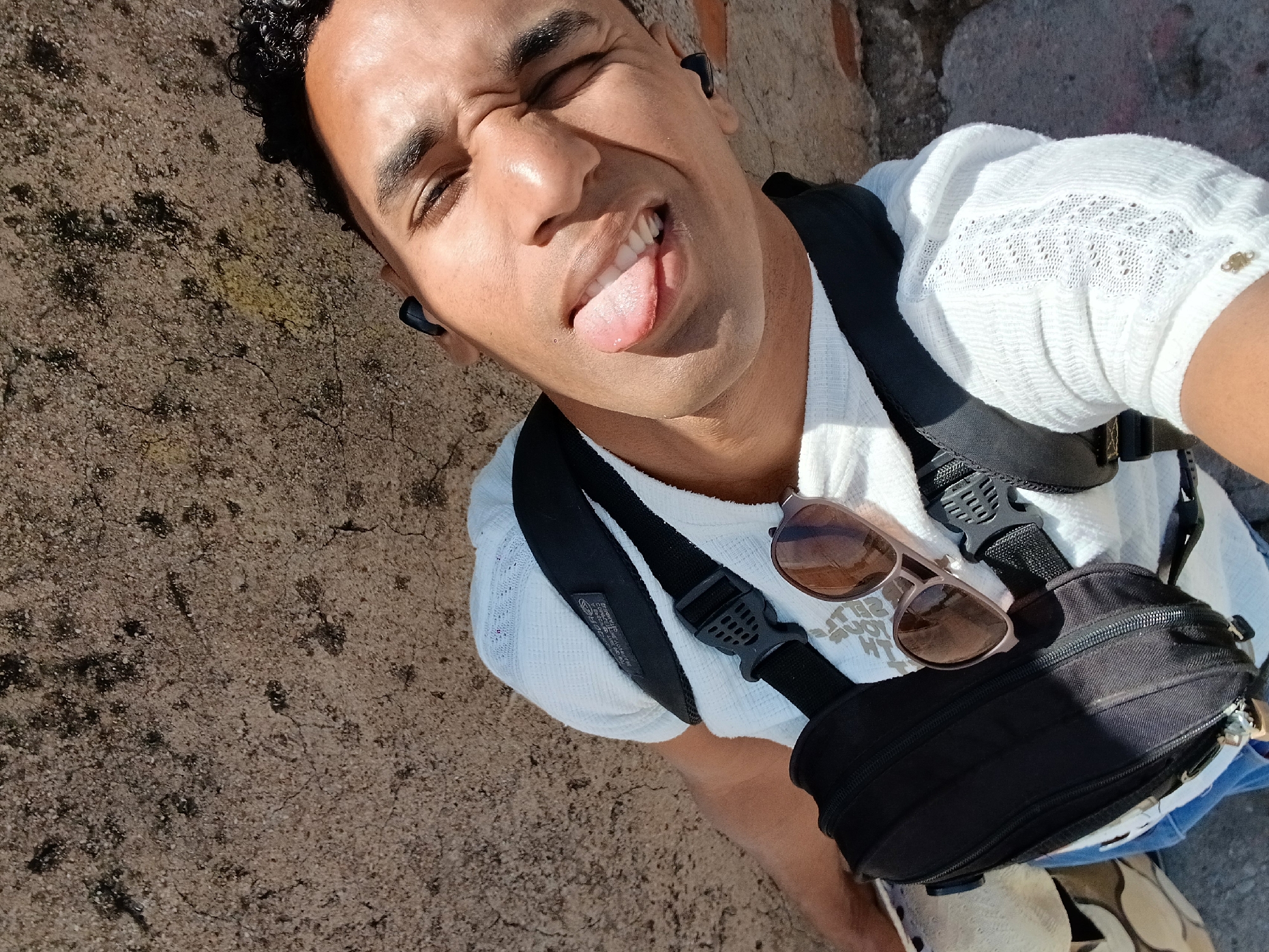 A man takes a selfie with his tongue sticking out. He wears a white shirt, a black fanny pack, and sunglasses. He wears a black earpiece in his left ear. The location is in Porlamar, Venezuela. The background is a concrete surface. The man is likely standing or sitting on the ground.
