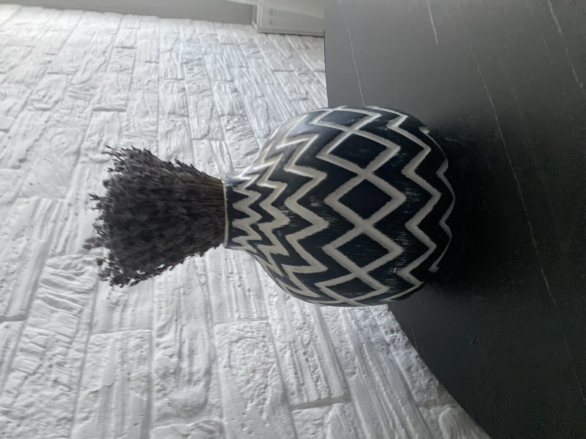 A vase filled with dried lavender sits on a black, textured table in front of a white, textured brick wall. The vase is round with a narrow neck and features a dark blue and white geometric pattern of zigzags and diamonds. The dried lavender is dark purple and bunches together at the top of the vase. The lighting is soft, casting a gentle shadow of the vase on the table. No people or actions are visible in the image. The location is not specified in the image or the provided context.