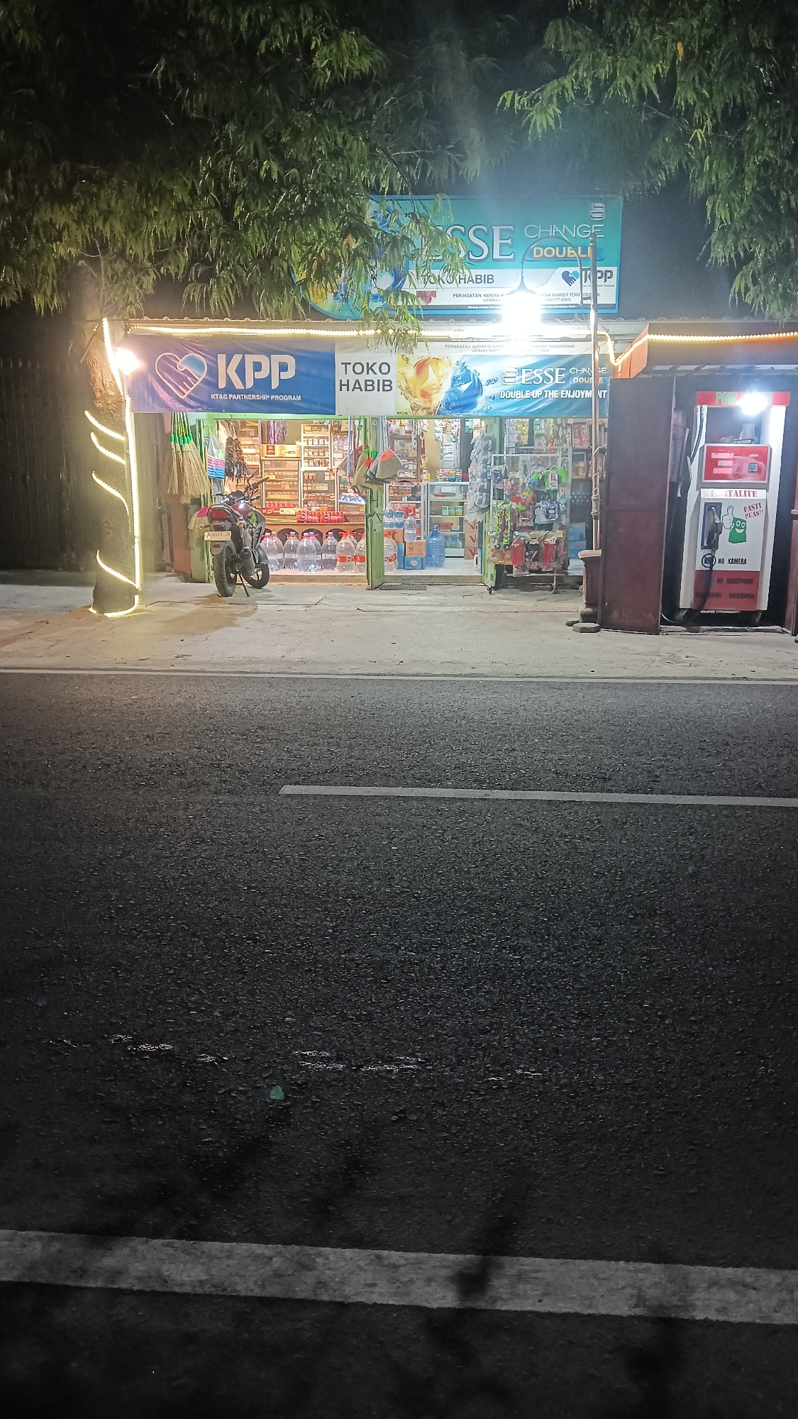 A nighttime exterior shot captures a roadside store in Sragen, Indonesia. The store, "TOKO HABIB," is well-lit and stocked with various goods, including bottled drinks. A motorcycle is parked in front of the store. The building features a large banner with "KPP KTAQ PARTNERSHIP PROGRAM" and another above with "ESSE CHANGE DOUBLE." To the right, a red and white gas pump stands, displaying the words "PASTE PLUS!!" and "NO KAMERA." The street in front of the store is paved and appears wet, with faint white lines marking the lane. Trees with dense foliage overhang the scene from above.