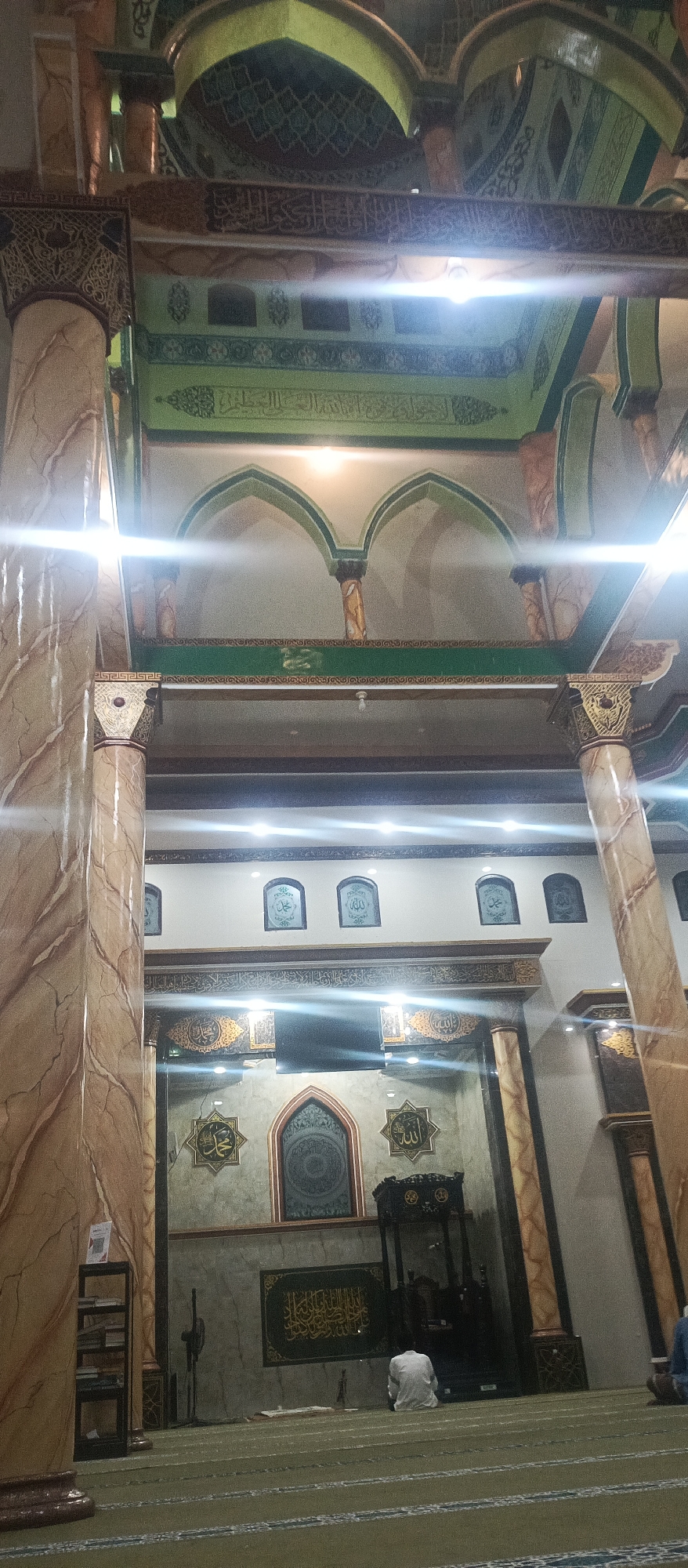 The image shows the interior of a mosque, likely located in Arjasa, Indonesia. The mosque features tall, marble-patterned columns that support arched structures and ornate ceilings decorated with intricate geometric patterns and Arabic calligraphy. There are several arched alcoves on the wall, each containing decorative Islamic calligraphy. In the center of the mosque, a dark wooden minbar (pulpit) is visible, and a man in white clothing is seated on the green carpeted floor in prayer. The floor is marked with lines indicating prayer rows. Various religious motifs and calligraphy are displayed on the walls, including star-shaped emblems with Arabic script. The lighting appears to be artificial, with bright lights creating some glare in the image.
