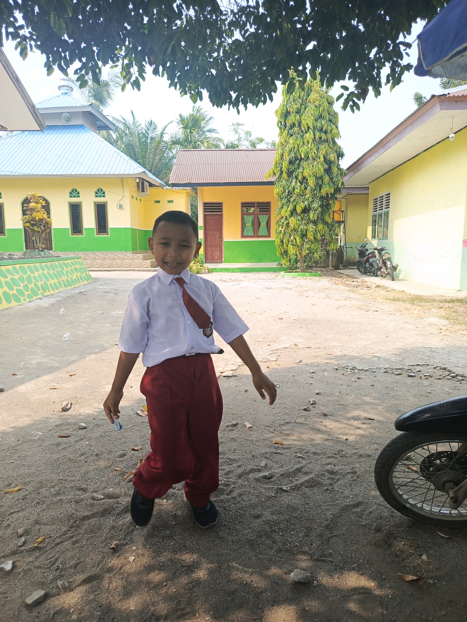 A young boy wearing a white short-sleeved school shirt with a red tie and red pants stands in the middle of a dusty outdoor area. The boy is smiling and looking towards the camera. Behind him, there are several buildings with yellow and green exteriors. One building has a blue roof. In the background, there are trees, including a tall, thin, green tree, and two motorcycles are parked near one of the buildings. The ground is covered in sand, gravel, and fallen leaves. The scene appears to be in front of a school building in Karya Dadi, Indonesia, as suggested by the context.