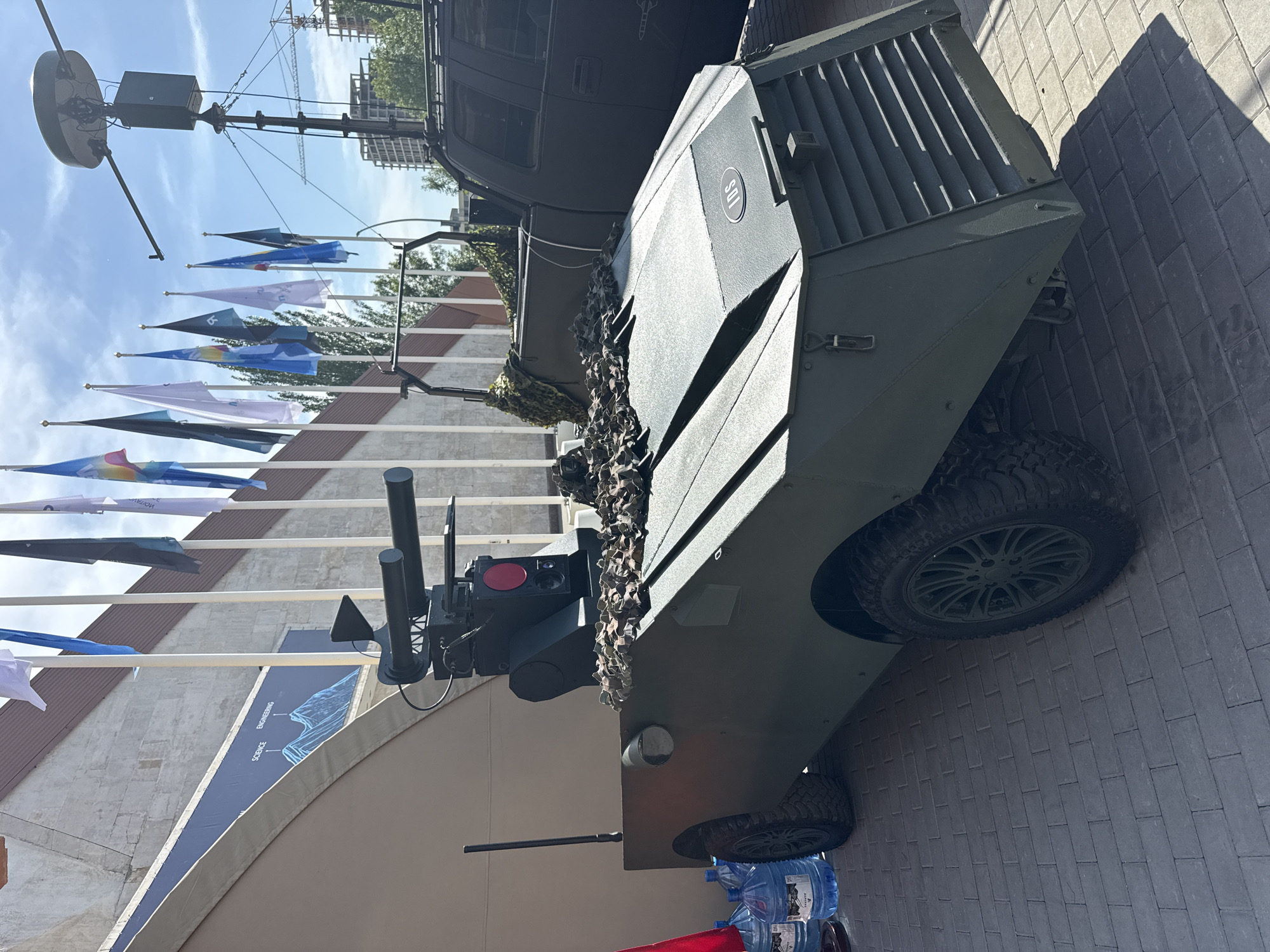 This image captures an outdoor exhibition scene on a bright, sunny day, with strong shadows indicating midday light. The primary subject is a dark olive-green military-style vehicle, likely an Unmanned Ground Vehicle (UGV) or a similar reconnaissance platform. It features rugged off-road tires and a mounted system at its front, which appears to be either a weapon or a sensor array, characterized by two cylindrical barrels, a prominent red circular element, and a small black conical sensor. Camouflage netting is draped over a section of the vehicle's chassis. A circular badge on the side of the vehicle clearly displays "SDS" and a possible number like "105" or "1OS".
In the background, a larger, dark military truck is partially visible. A tall mast equipped with a large circular antenna (possibly a satellite dish) and other communication gear extends upwards into the clear blue sky. A long row of poles displays numerous colorful, triangular flags or pennants, some bearing discernible text such as a stylized "R" and the reversed word "VOYAGE". Behind these flags, a building with a light-colored, possibly concrete, facade and reddish-brown roof elements can be seen. A large, dark blue banner with white text that reads "SCIENCE" and "ENGINEERING" is also part of the display. The ground is paved with interlocking bricks. Stacked near the front left wheel of the main vehicle are several large blue plastic water bottles, some with "WATER" visible on their labels. The scene suggests a defense technology exhibition or a public display of military equipment.